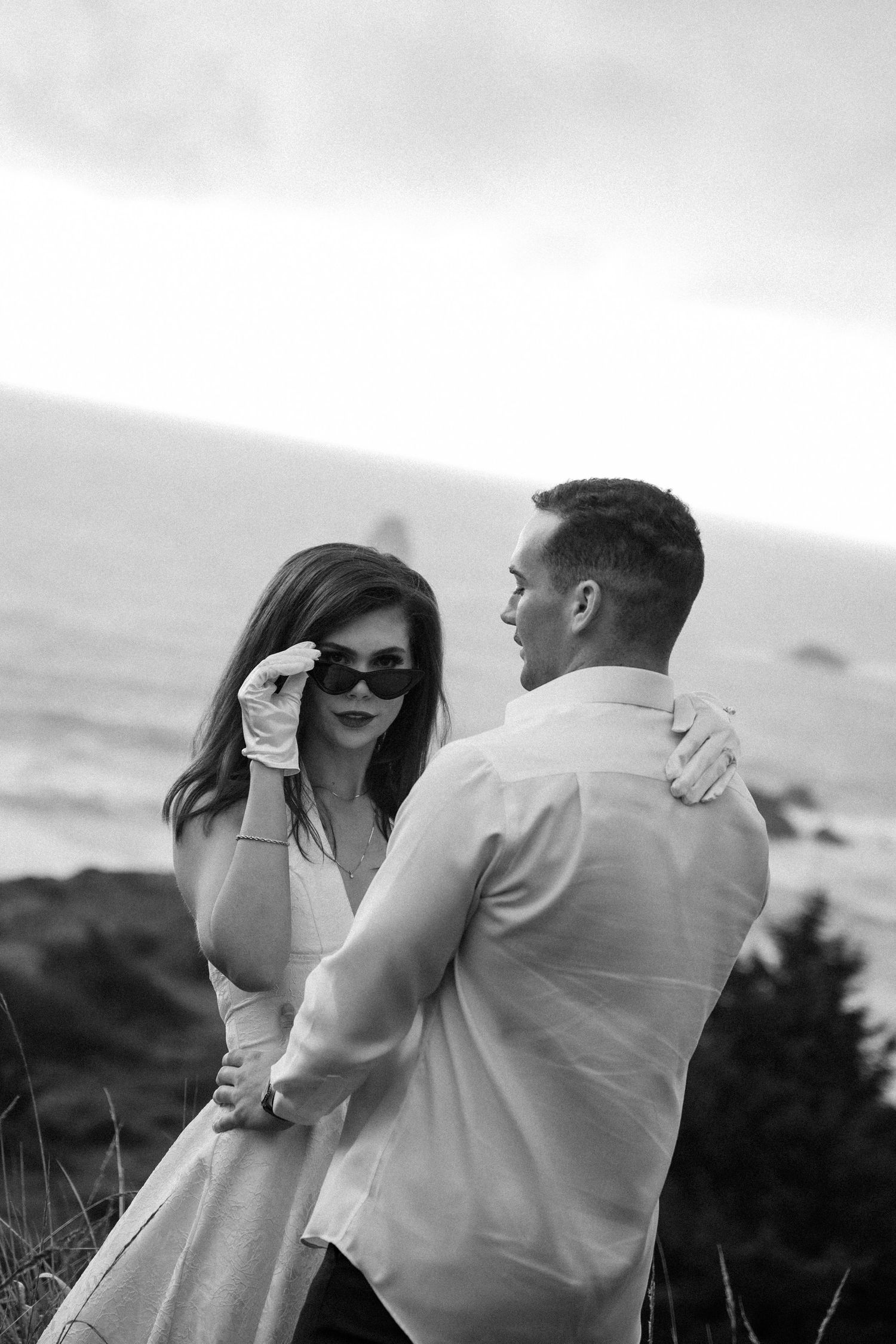 A couple shares an intimate moment on a coastal overlook in black and white.