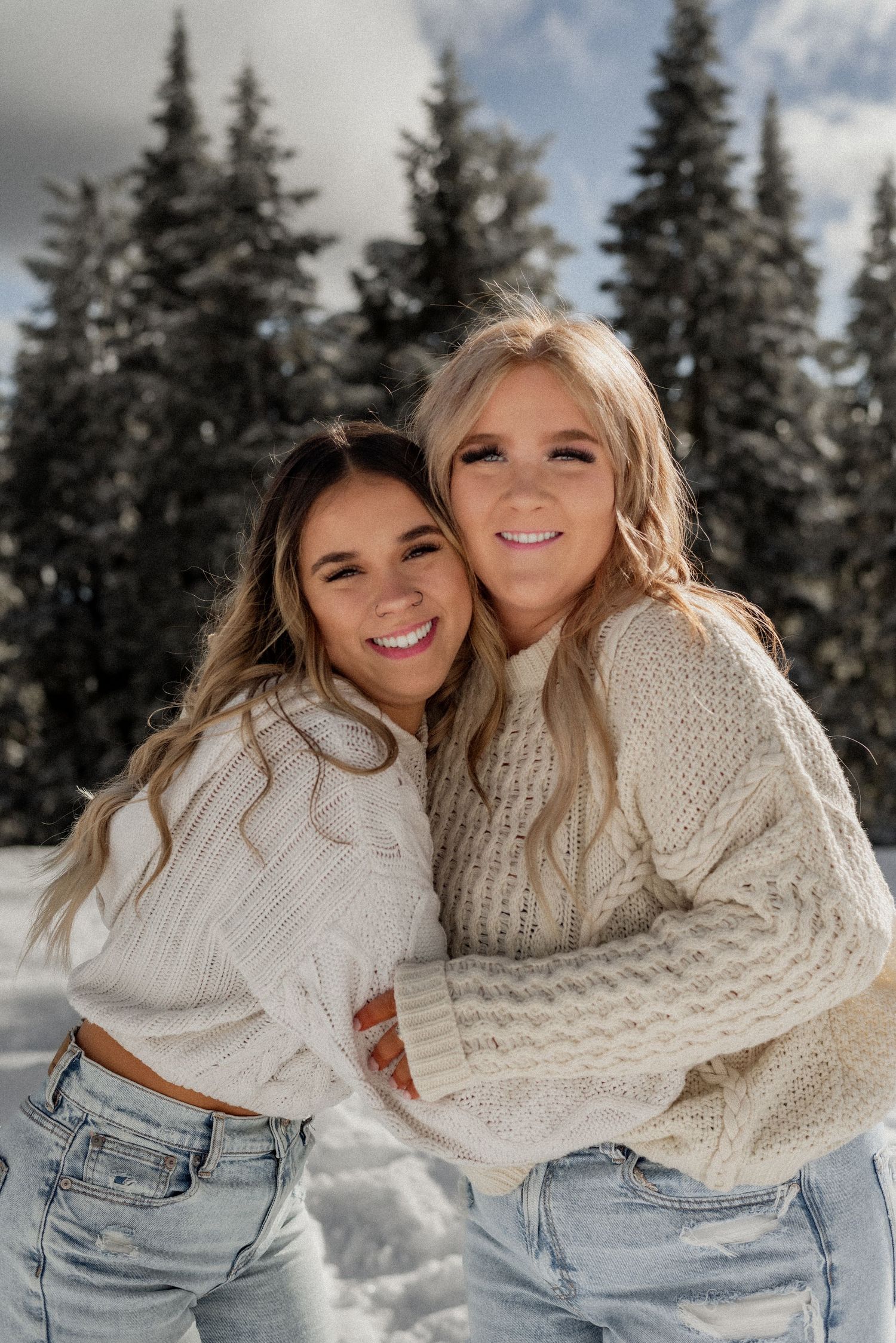Winter photoshoot of two friends in cream sweaters and jeans against snowy forest backdrop.