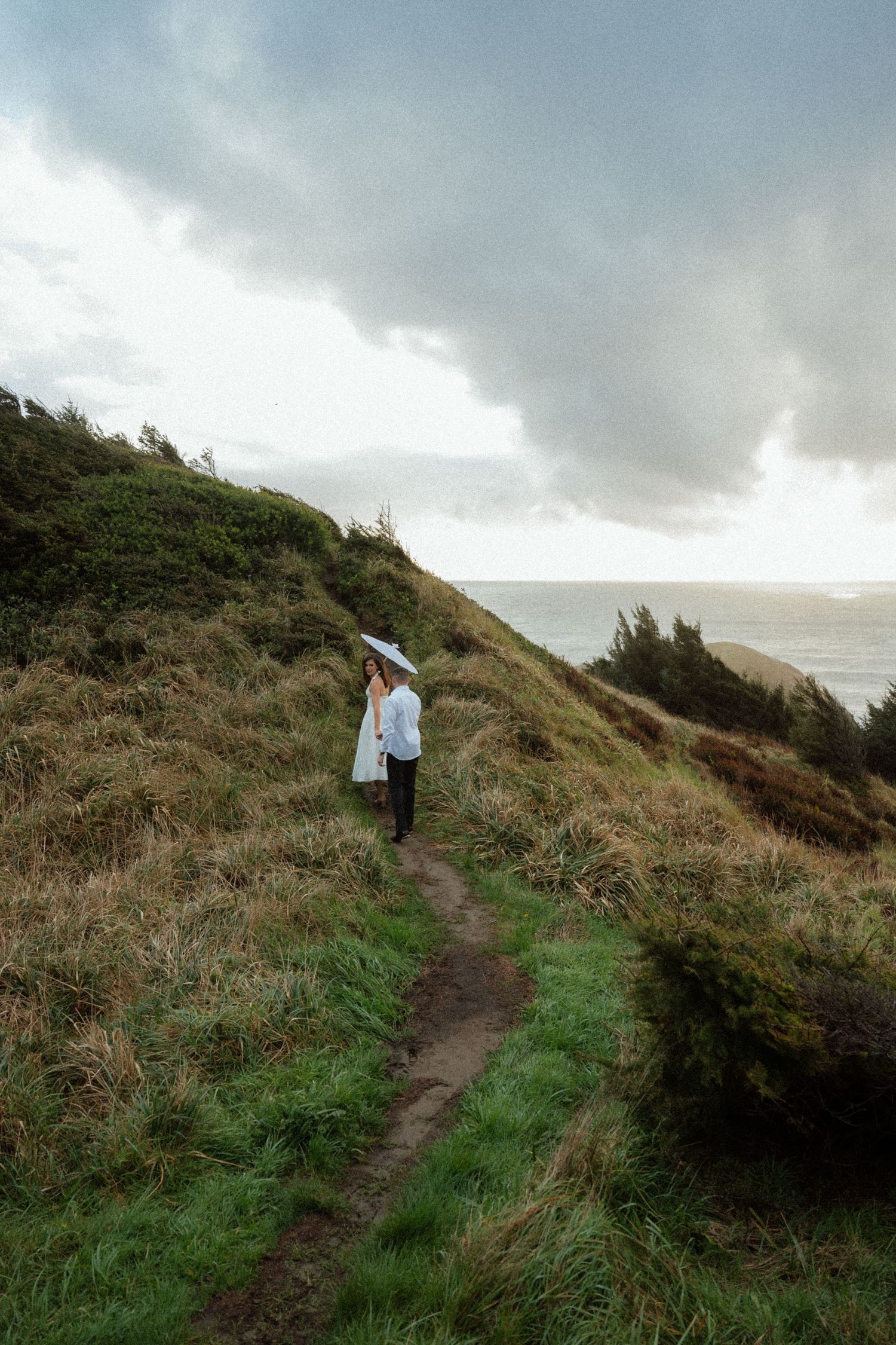 A couple walks hand in hand down a coastal trail under cloudy skies.