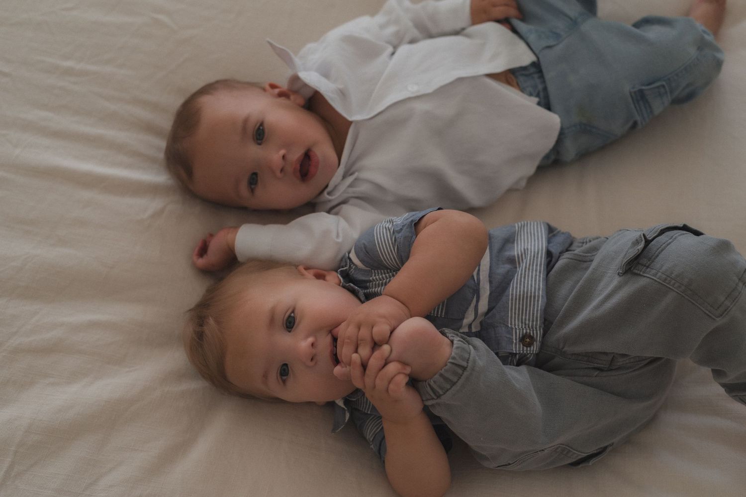 Baby in casual outfit lies on white bedding while playing and smiling.