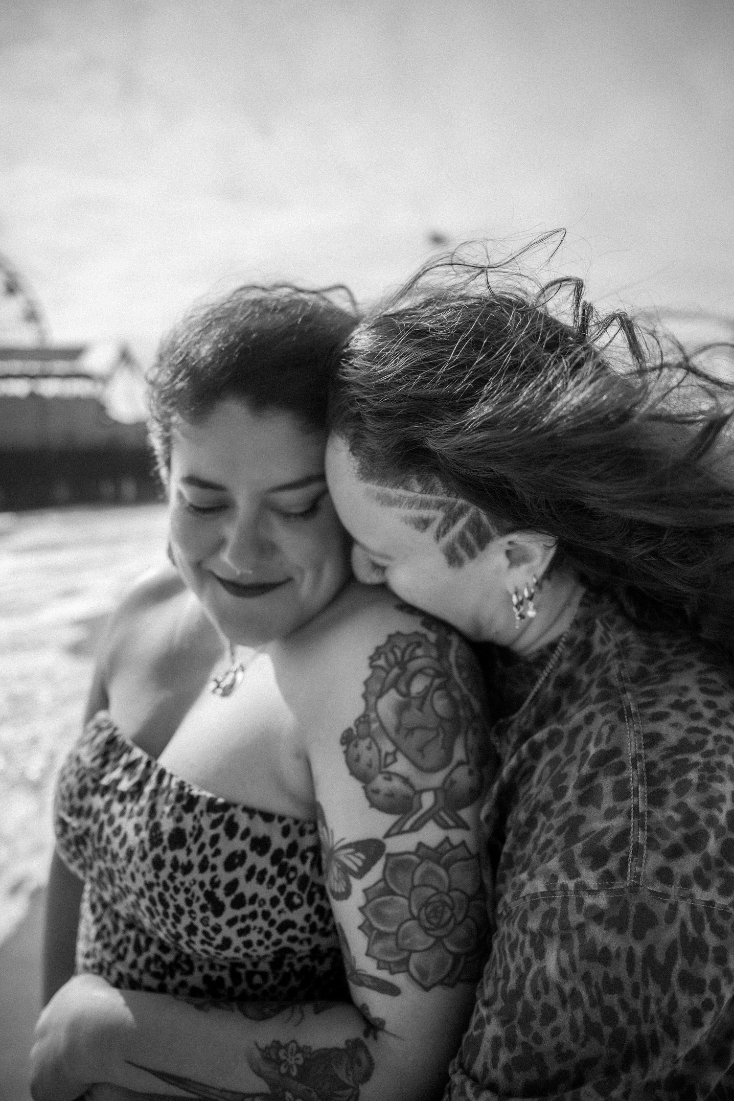 An intimate black and white portrait of two people with visible tattoos sharing a tender moment at the beach.