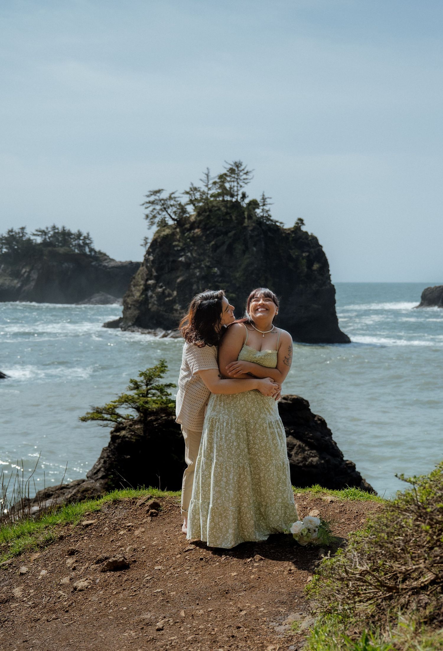 Two people in summer dresses share a moment on scenic coastal cliffs.