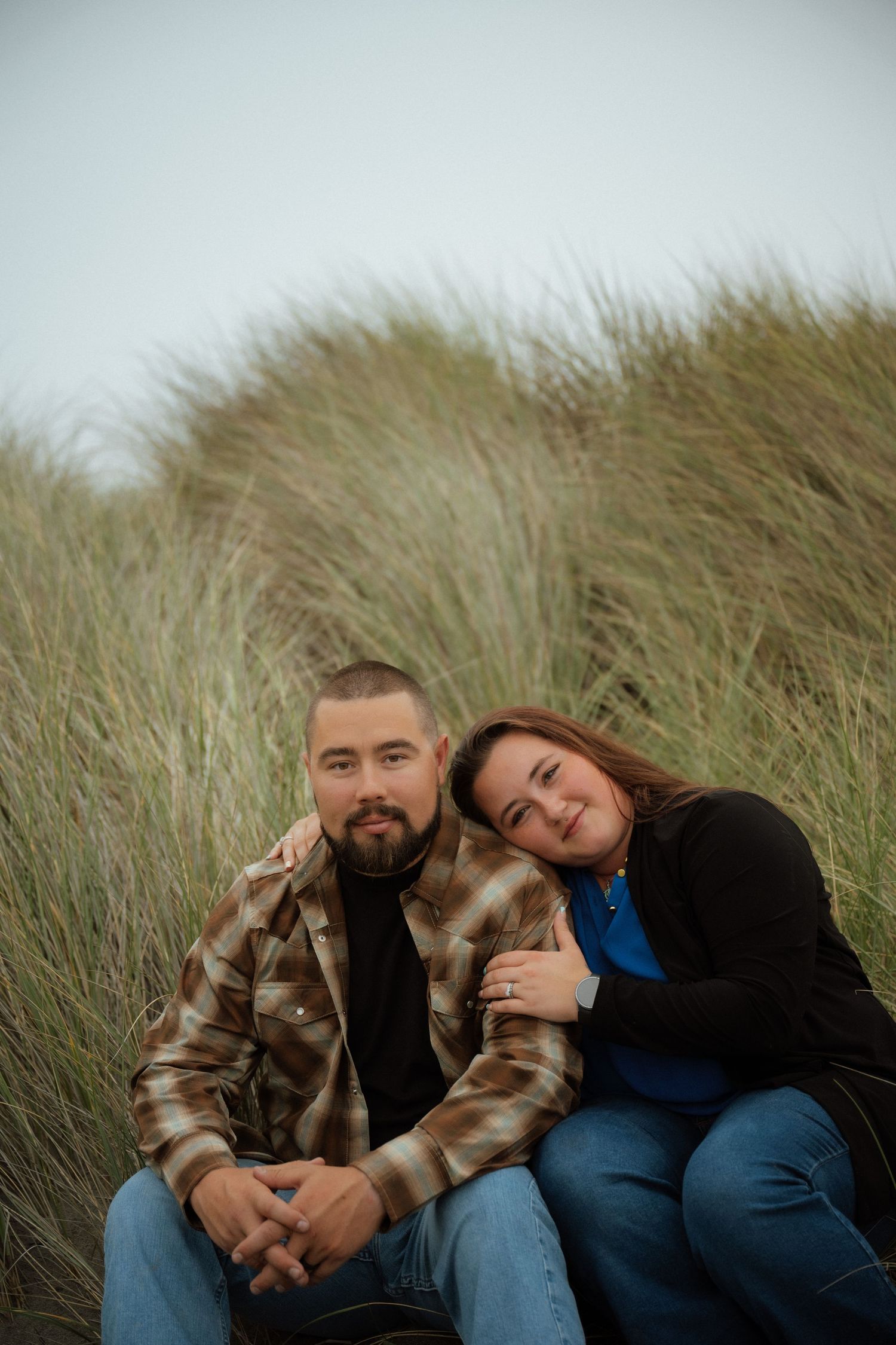 A couple sits together in tall beach grass during an overcast day.