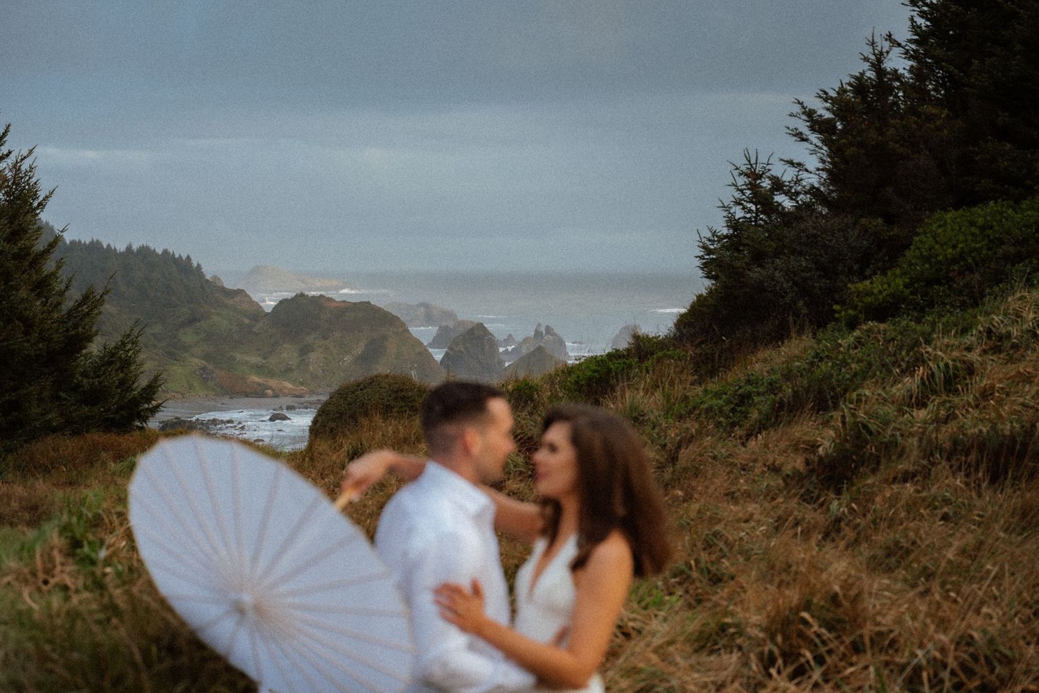 Two people share a romantic embrace on a misty coastal cliff with evergreen trees.