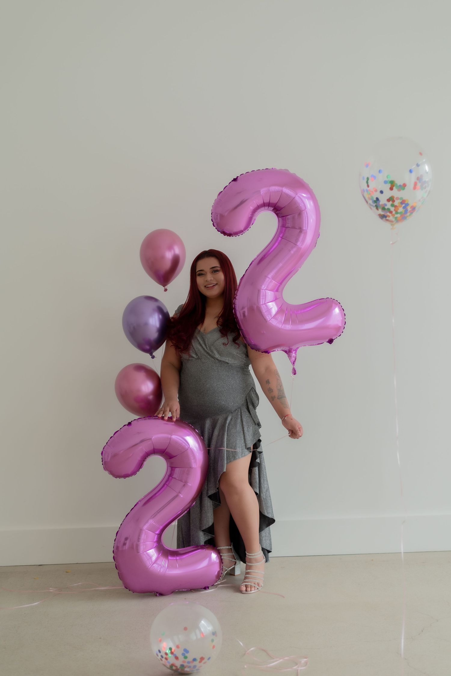 Woman in gray dress poses with pink balloons and confetti in bright studio setting.