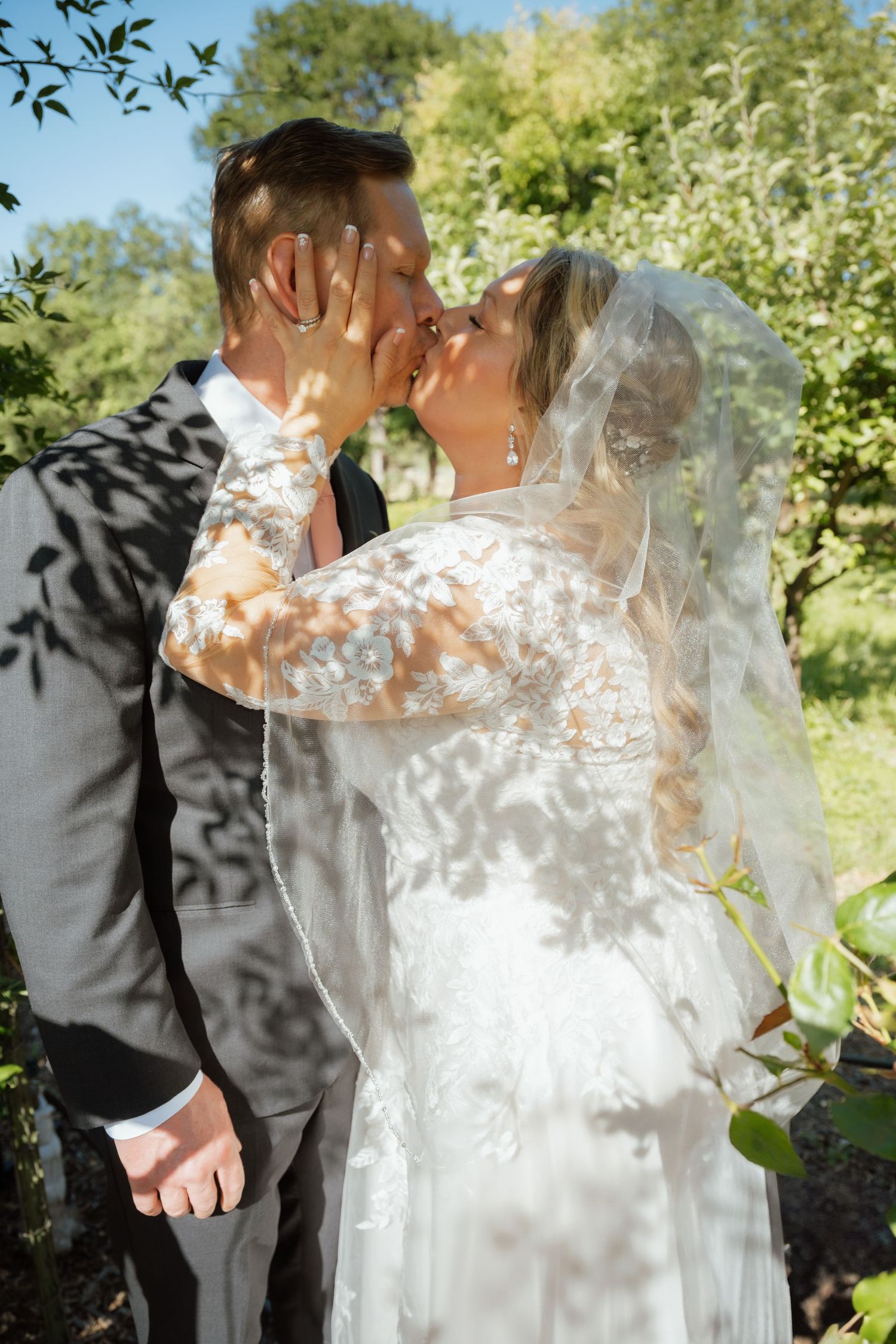 A romantic wedding kiss captured through delicate shadows of leaves and sunlight.
