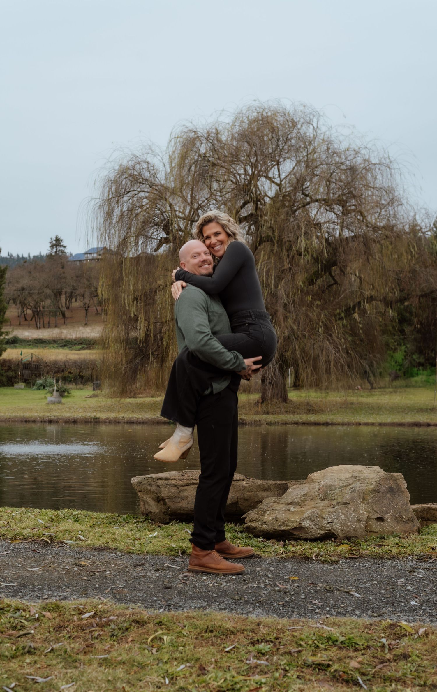 Couple posing playfully by lake with weeping willow trees in background.