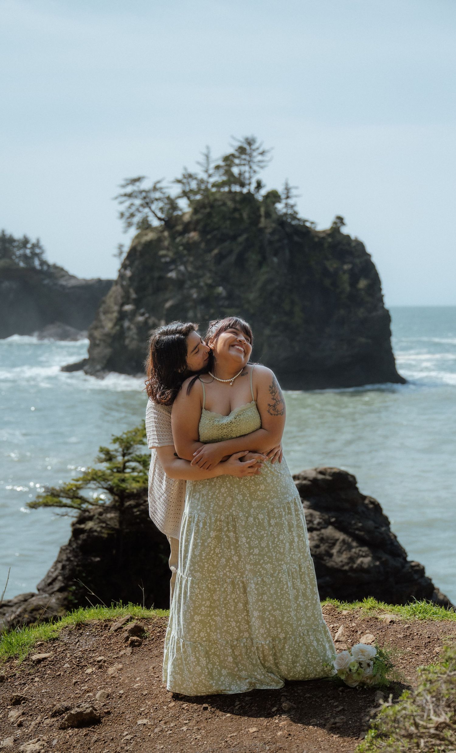 Two people share an affectionate moment against a backdrop of coastal cliffs and ocean waves.