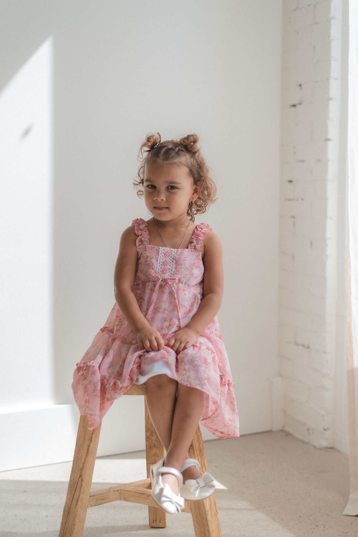 Small child in pink floral dress sits happily on wooden stool by sunlit window.