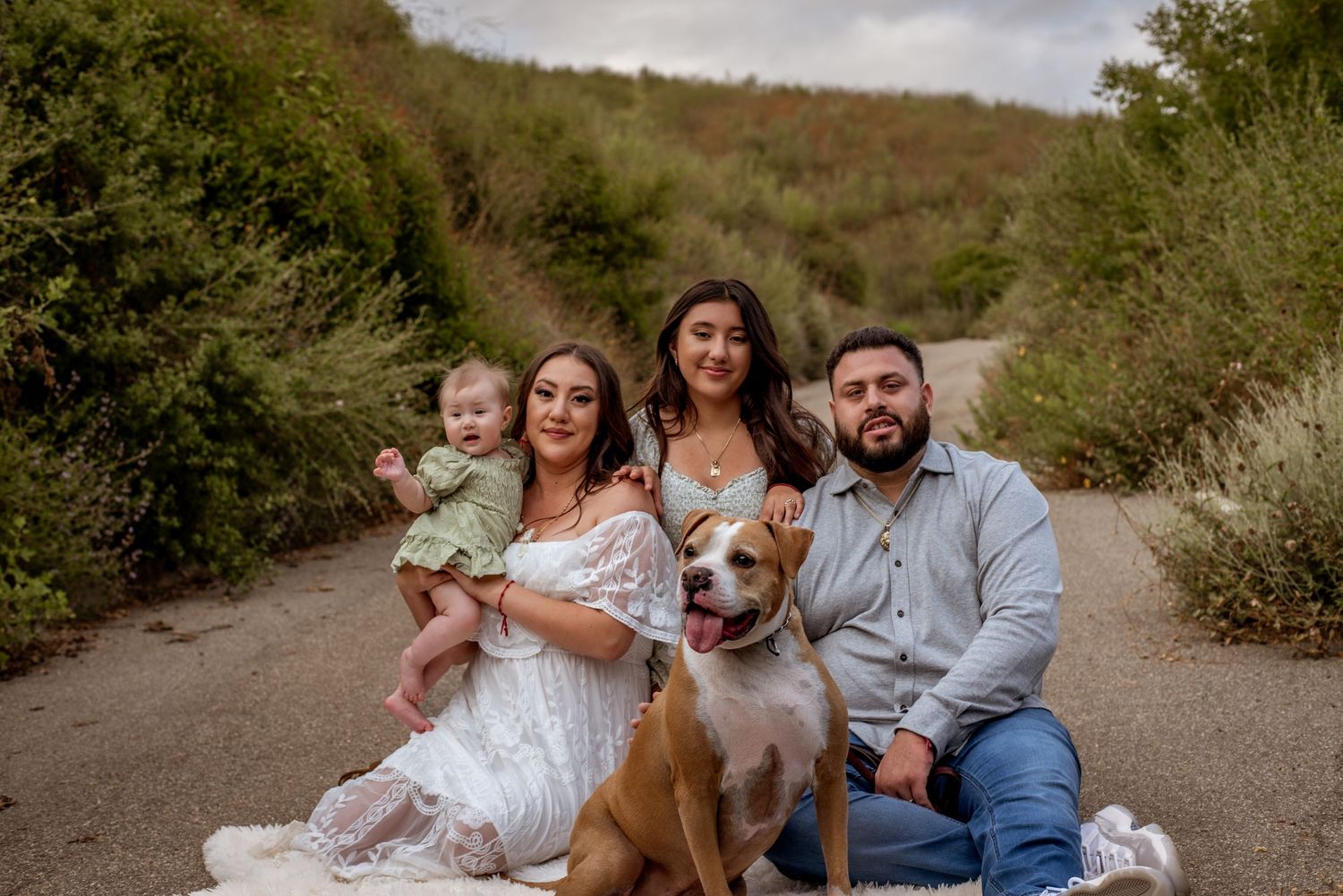 A family poses together with their brown dog on a dirt path surrounded by greenery.