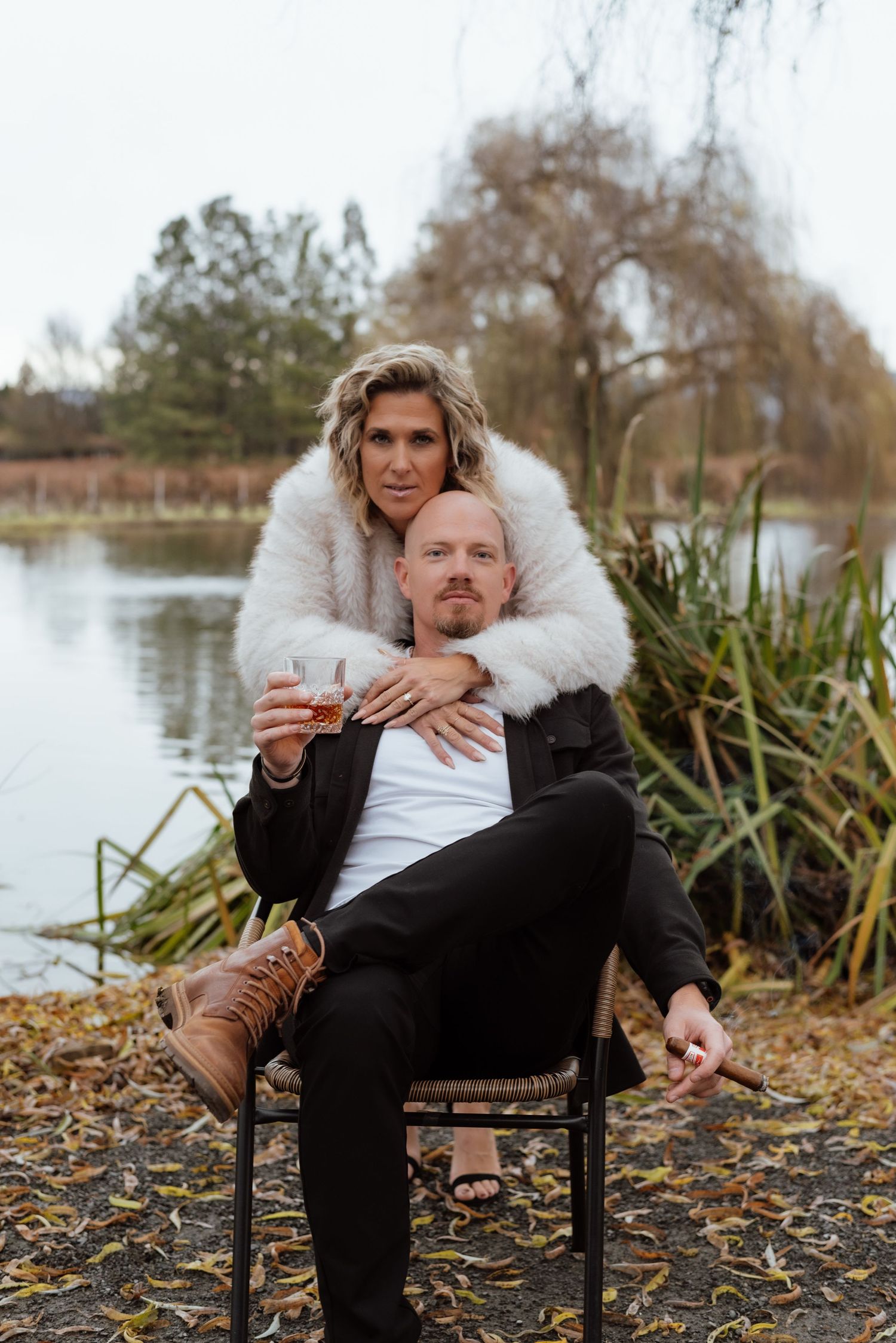 Couple relaxing on chair by pond, with person wearing white fuzzy coat embracing from behind.