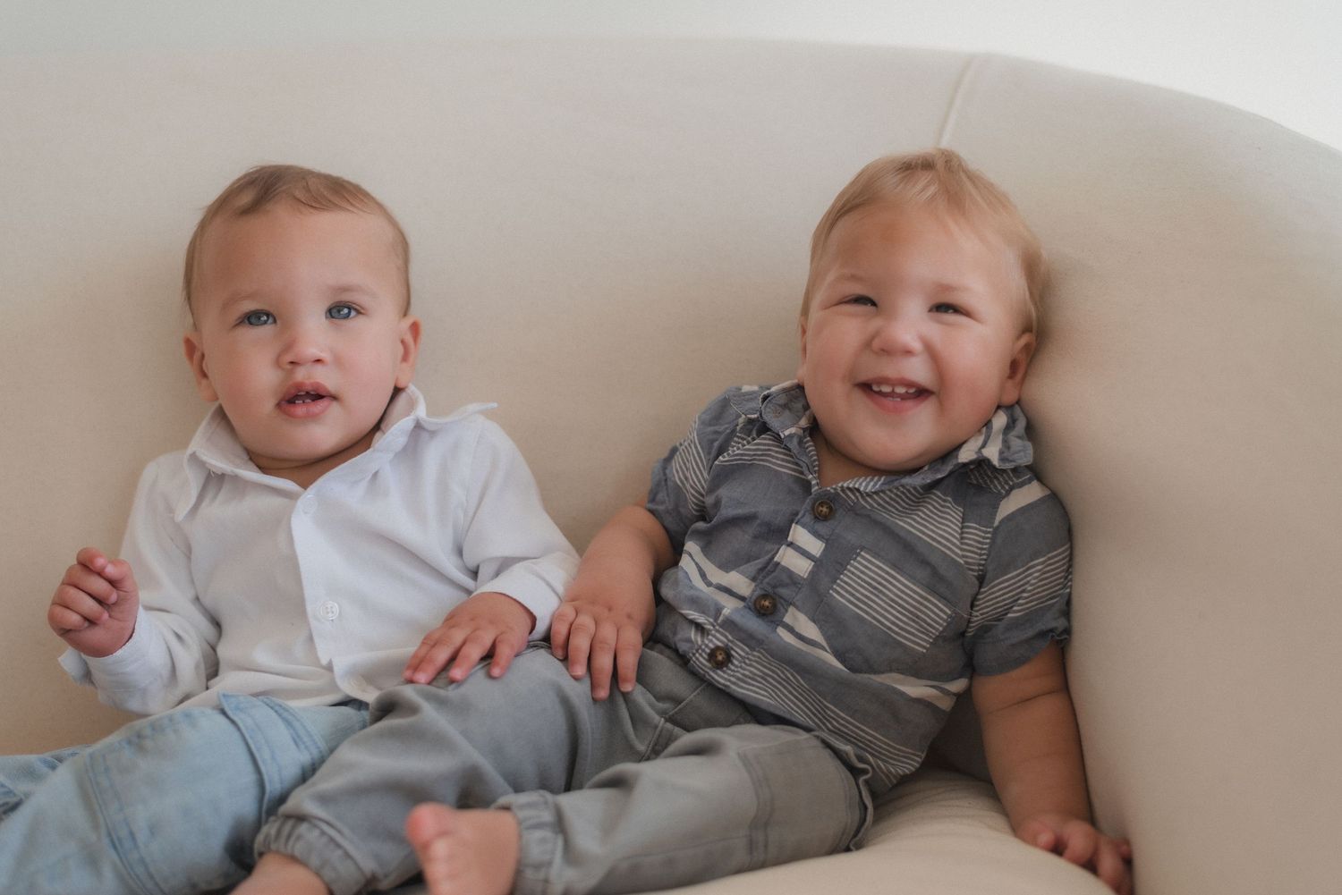 Two babies in matching gray outfits sitting together on white furniture.