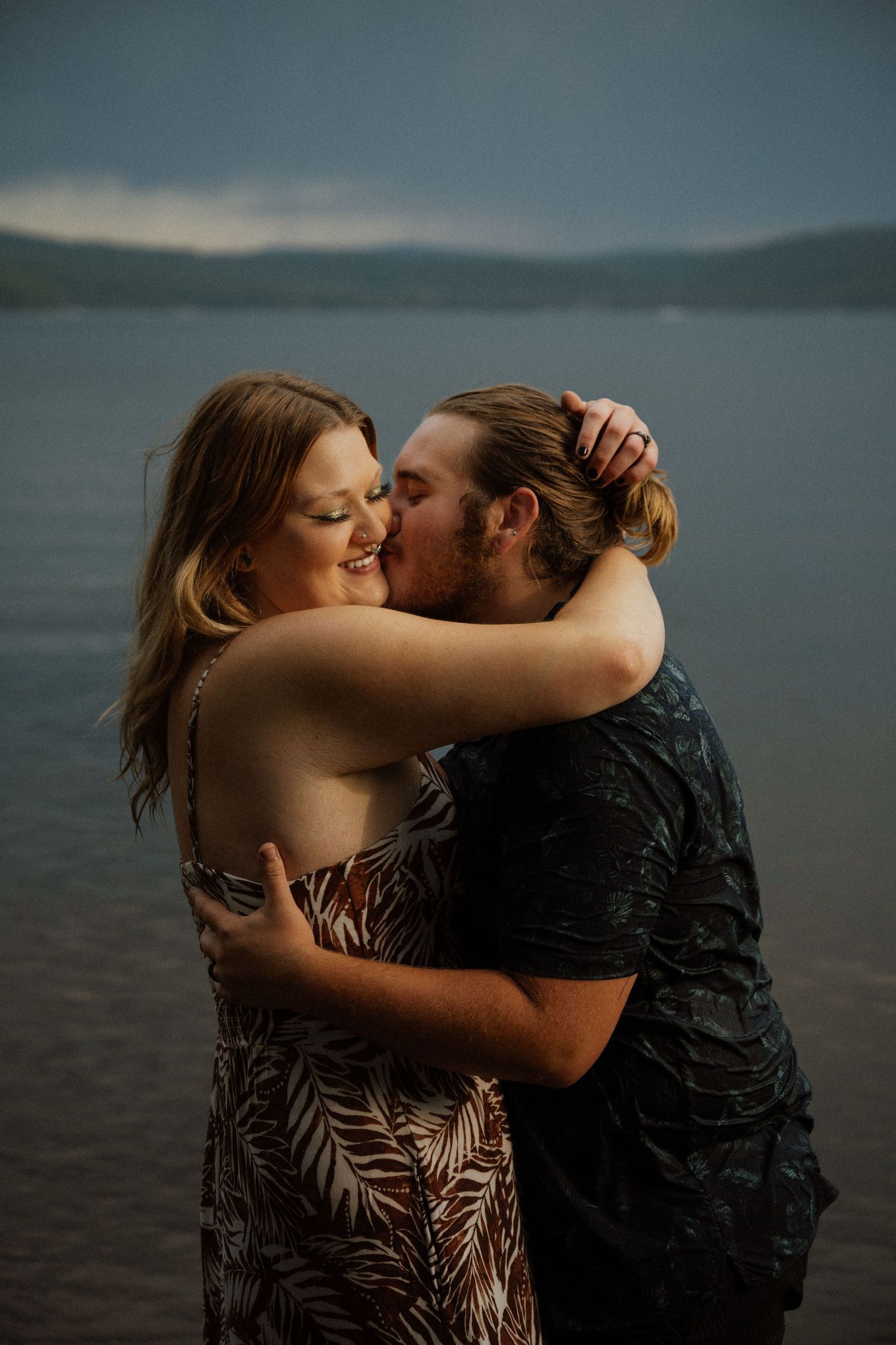 Romantic couple embracing on a moody lakeside with mountains in the background.