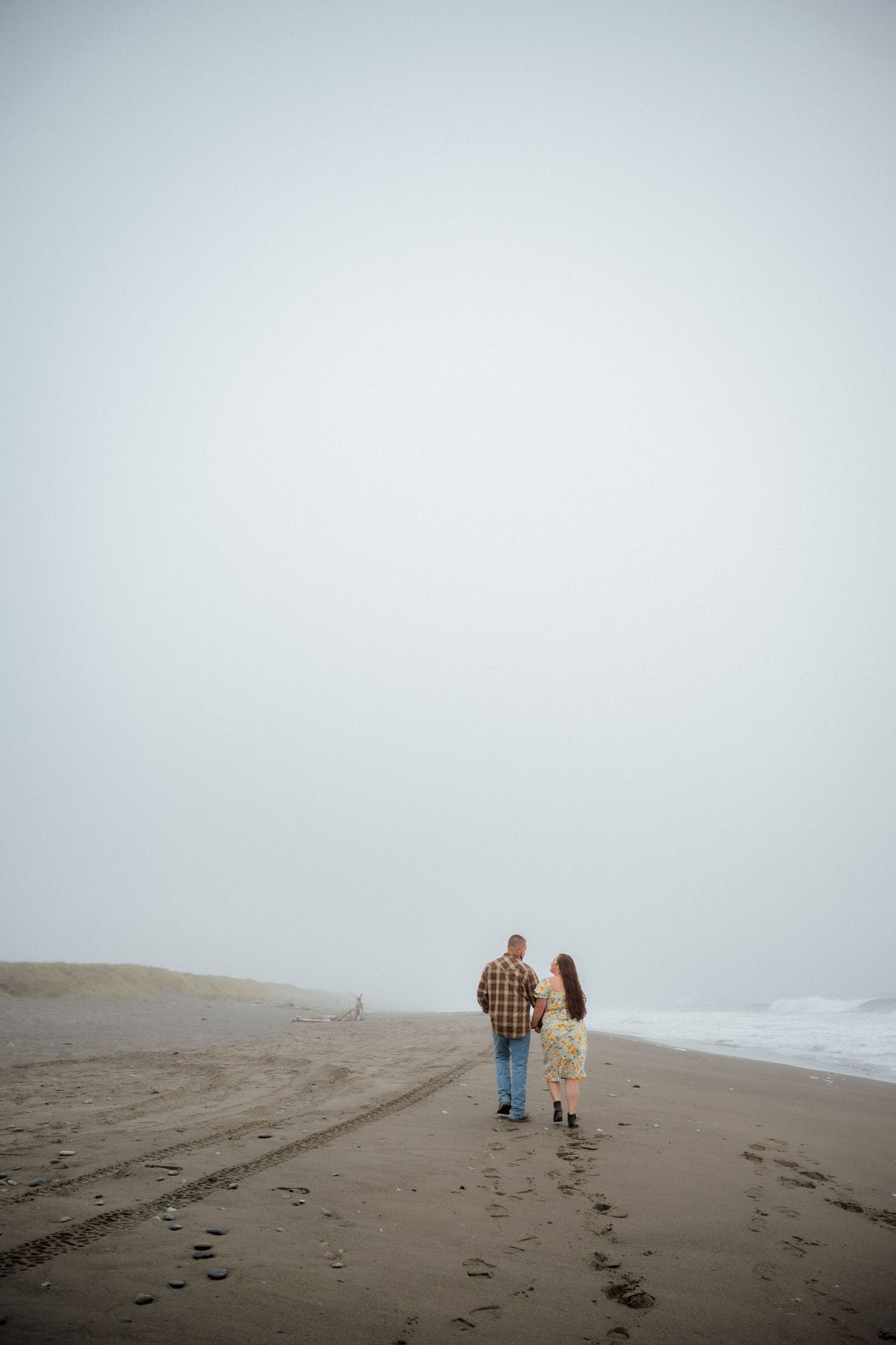 A couple walks along a misty beach leaving footprints in the sand on an overcast day.