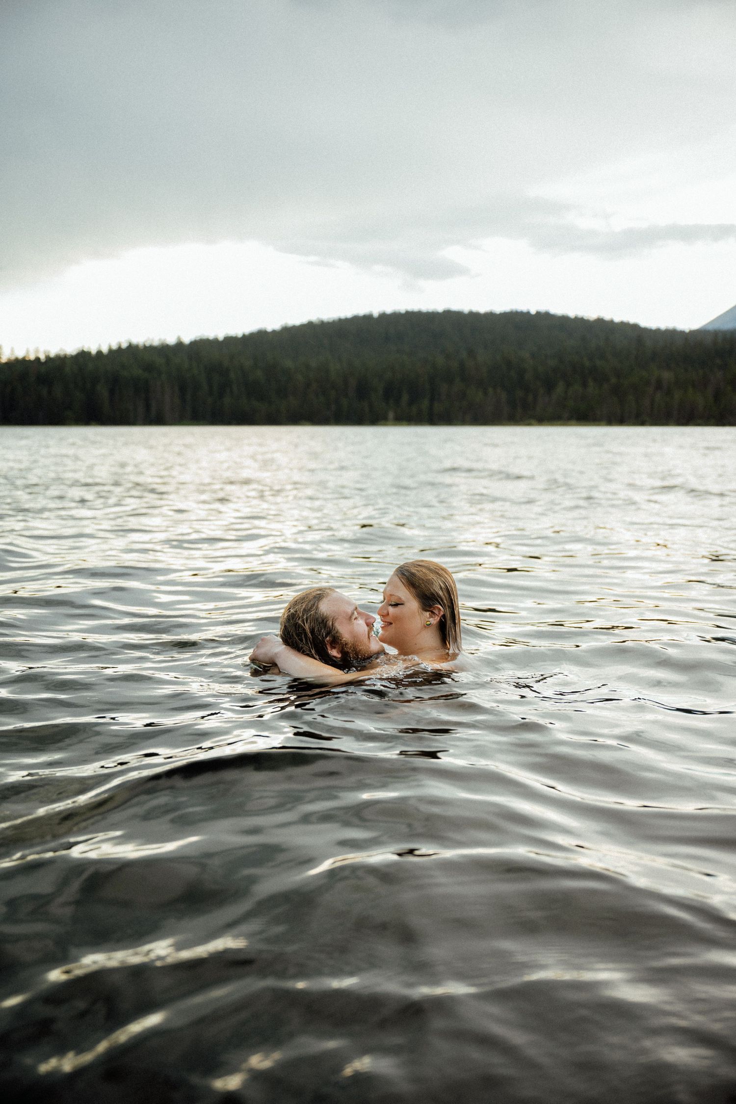 Two people floating close together in dark lake waters with mountains in the background.