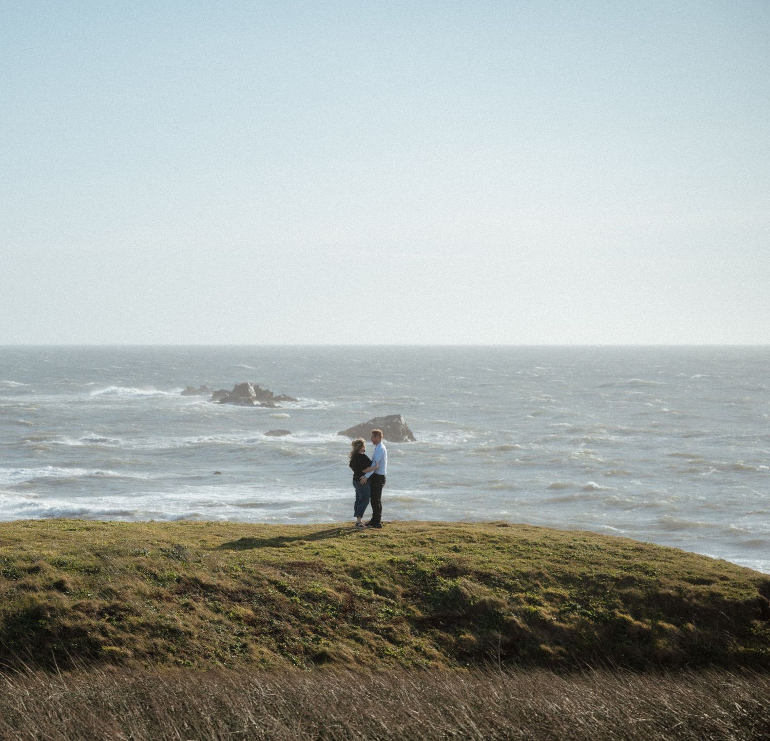 Two figures stand on a windswept coastal cliff overlooking a dramatic ocean landscape.