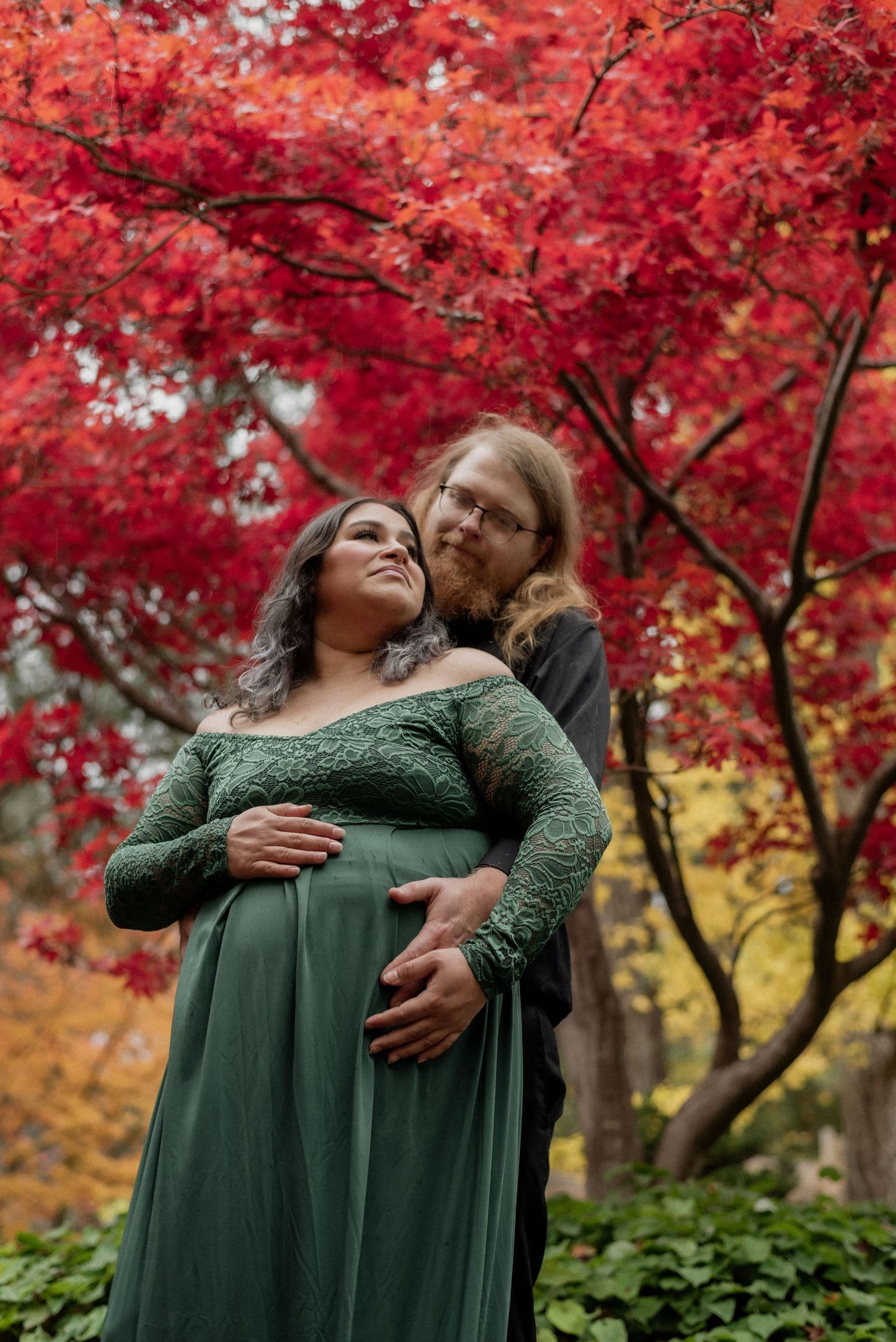 A romantic maternity photo featuring a green dress against vibrant red maple leaves.