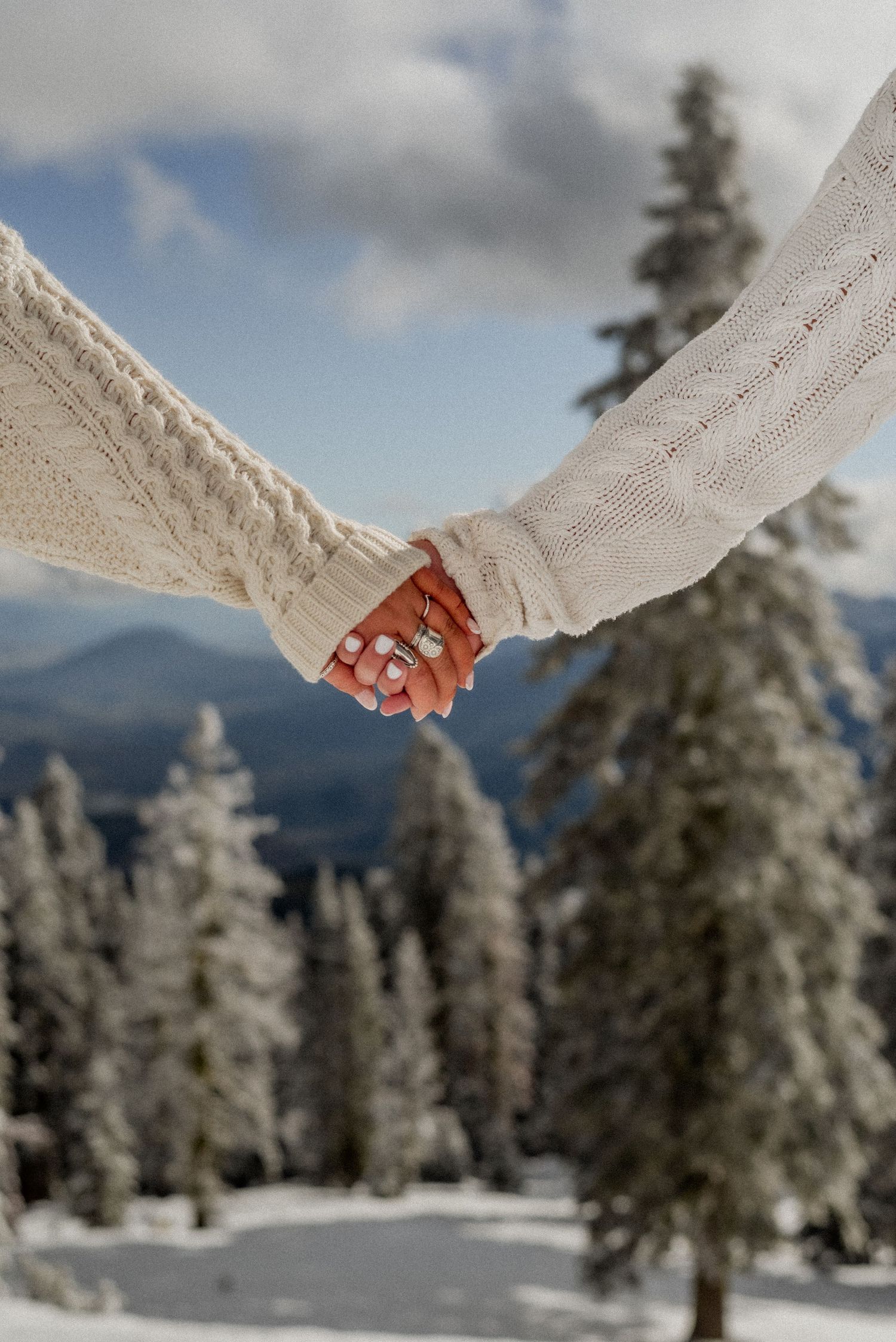 Two people in white knit sweaters hold hands against a snowy mountain landscape with evergreen trees.