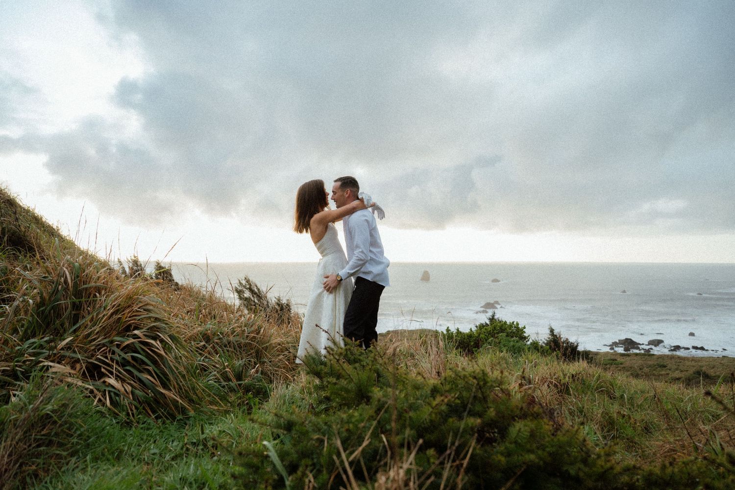 A couple stands together on a grassy coastal bluff overlooking the ocean.