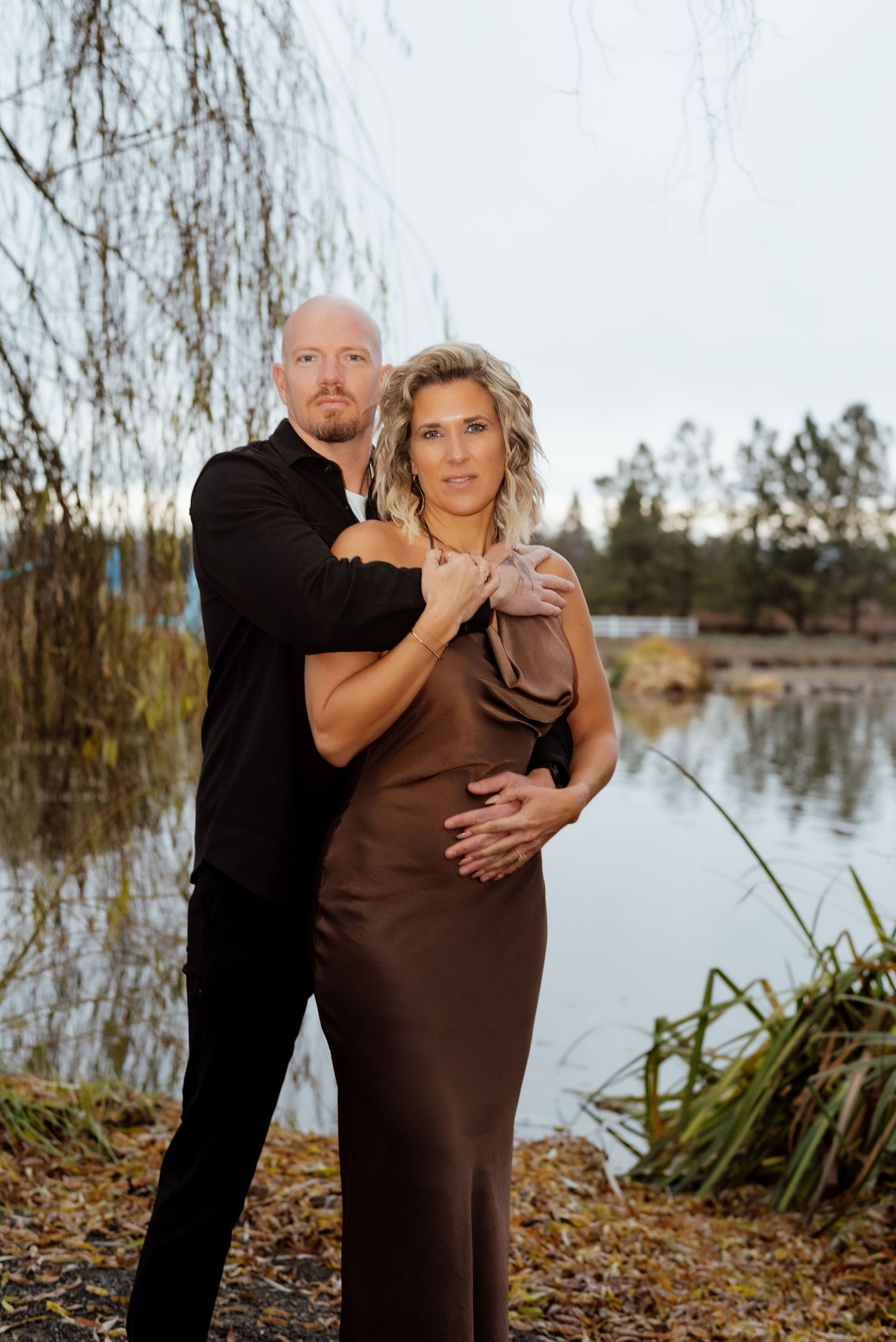 Romantic maternity photoshoot by a lake with couple in brown dress and black attire among fallen leaves.