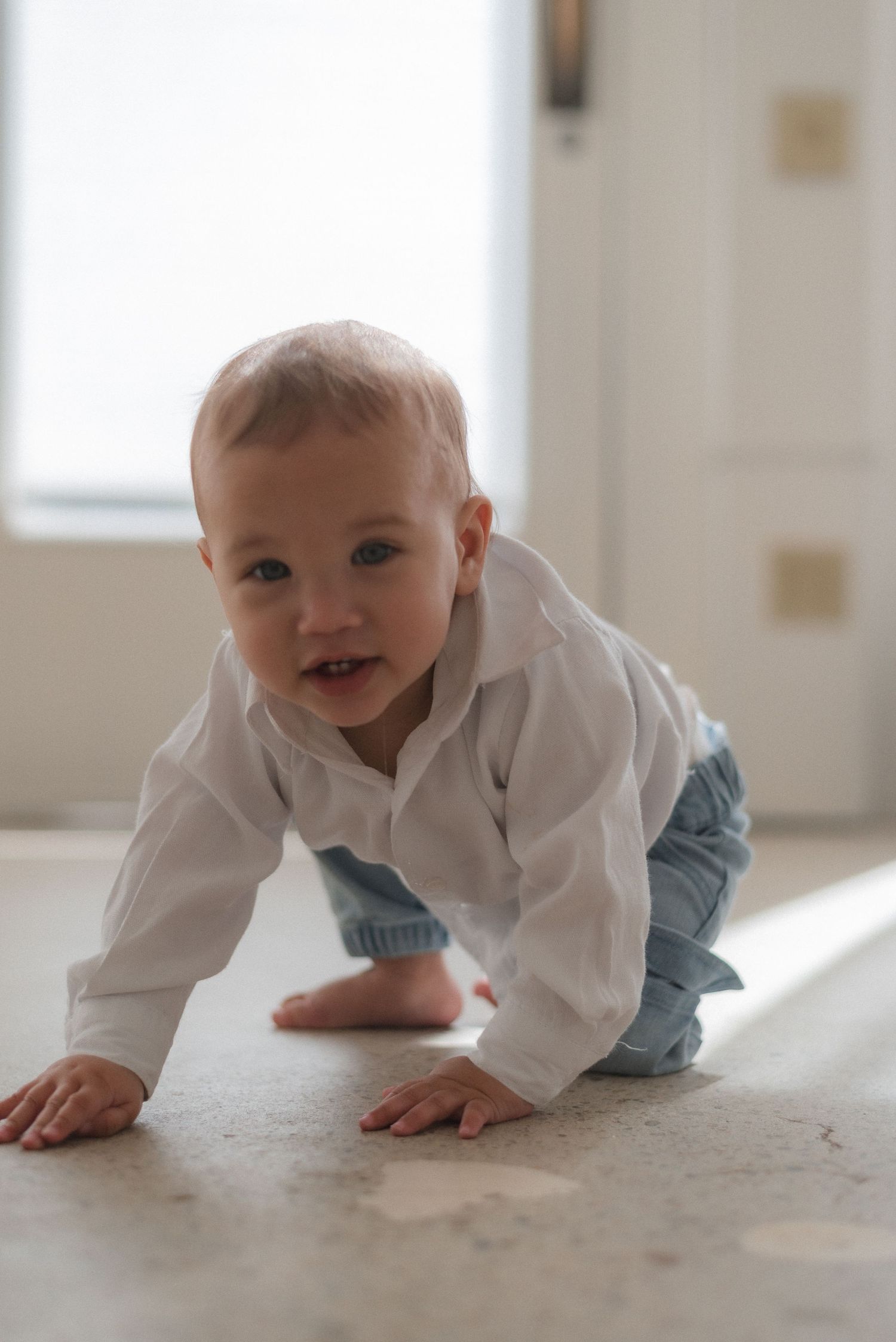 Baby in white shirt and blue pants crawls on light colored floor in sunny room.