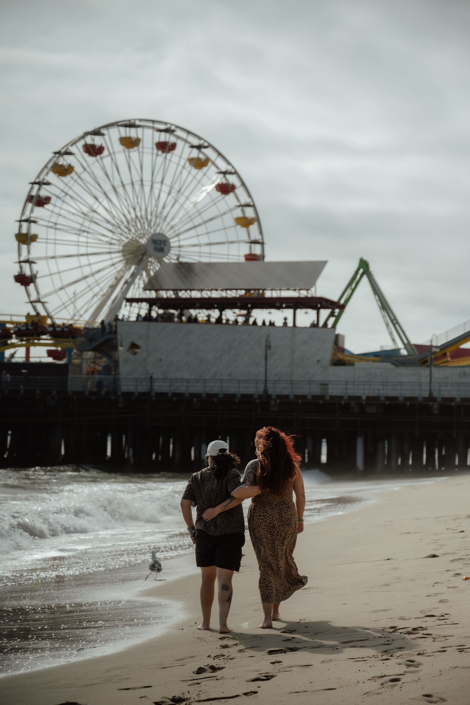 A couple strolls along the beach near a pier with a prominent Ferris wheel against an overcast sky.