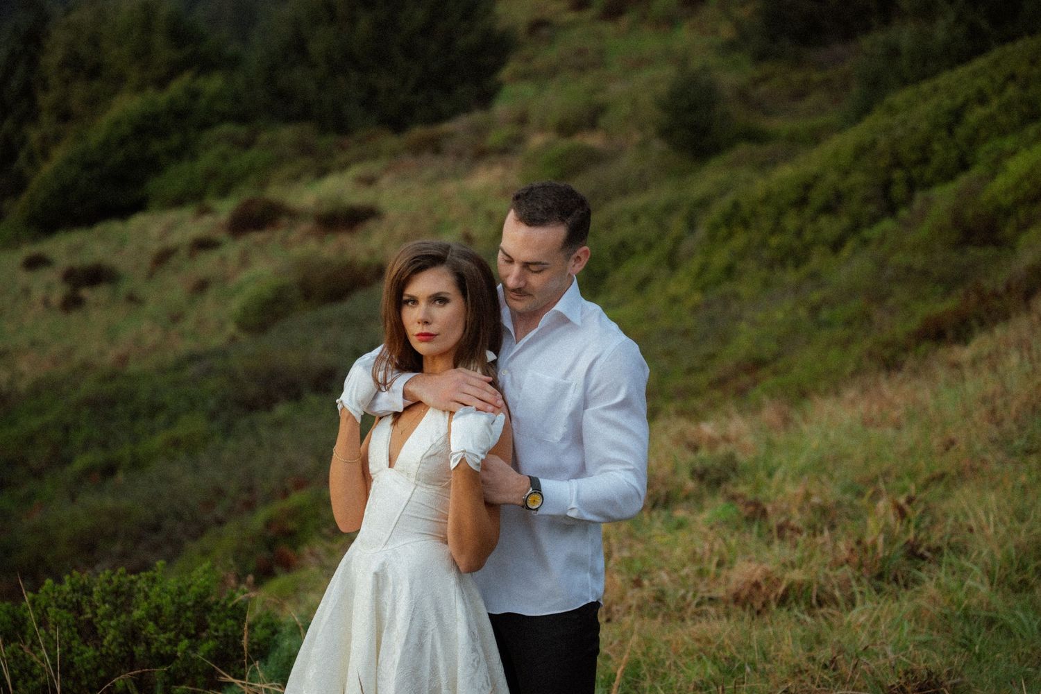A couple embraces on a scenic hillside overlooking the ocean under moody skies.