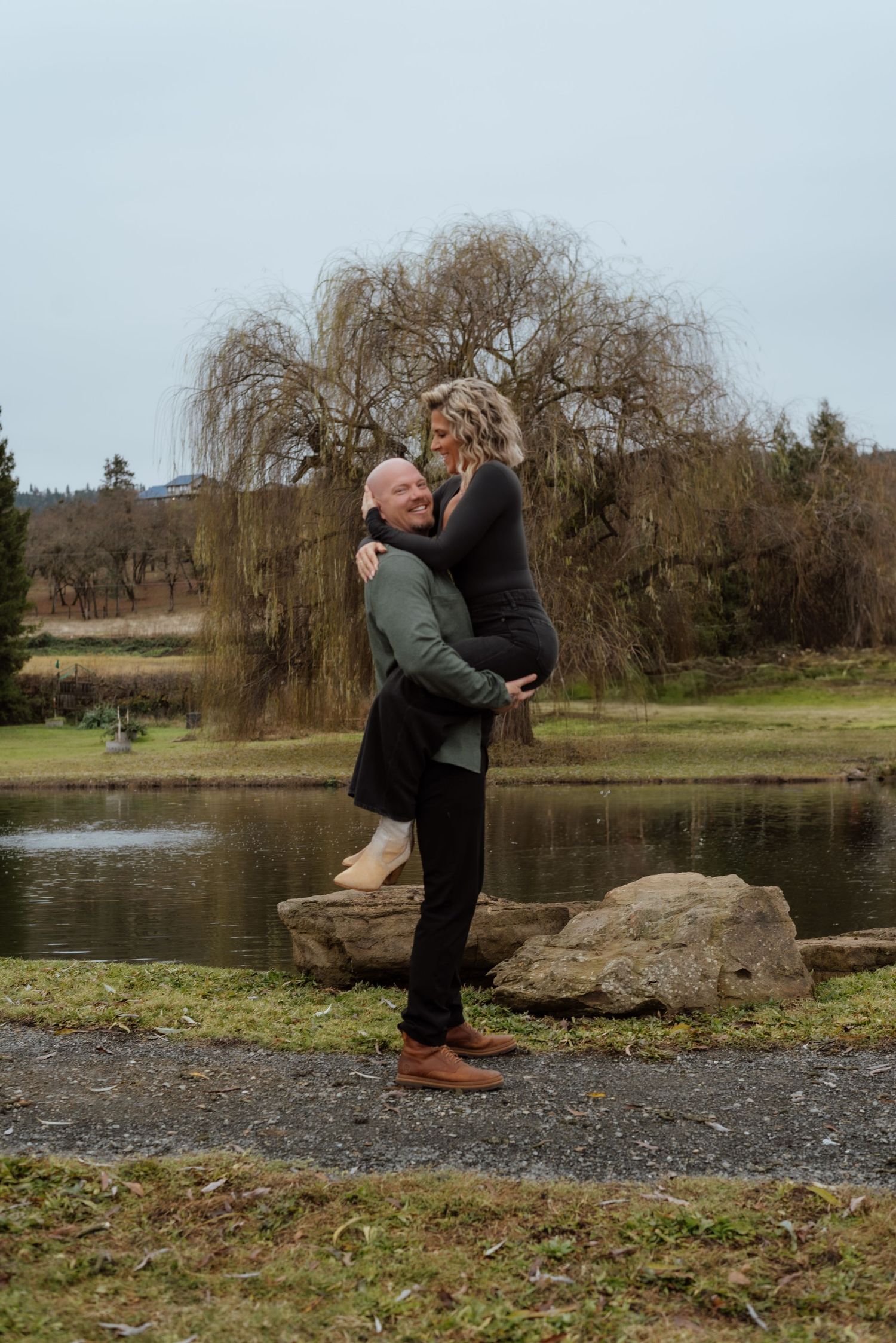 Romantic couple embraces by a pond with a weeping willow tree in the background on an overcast day.