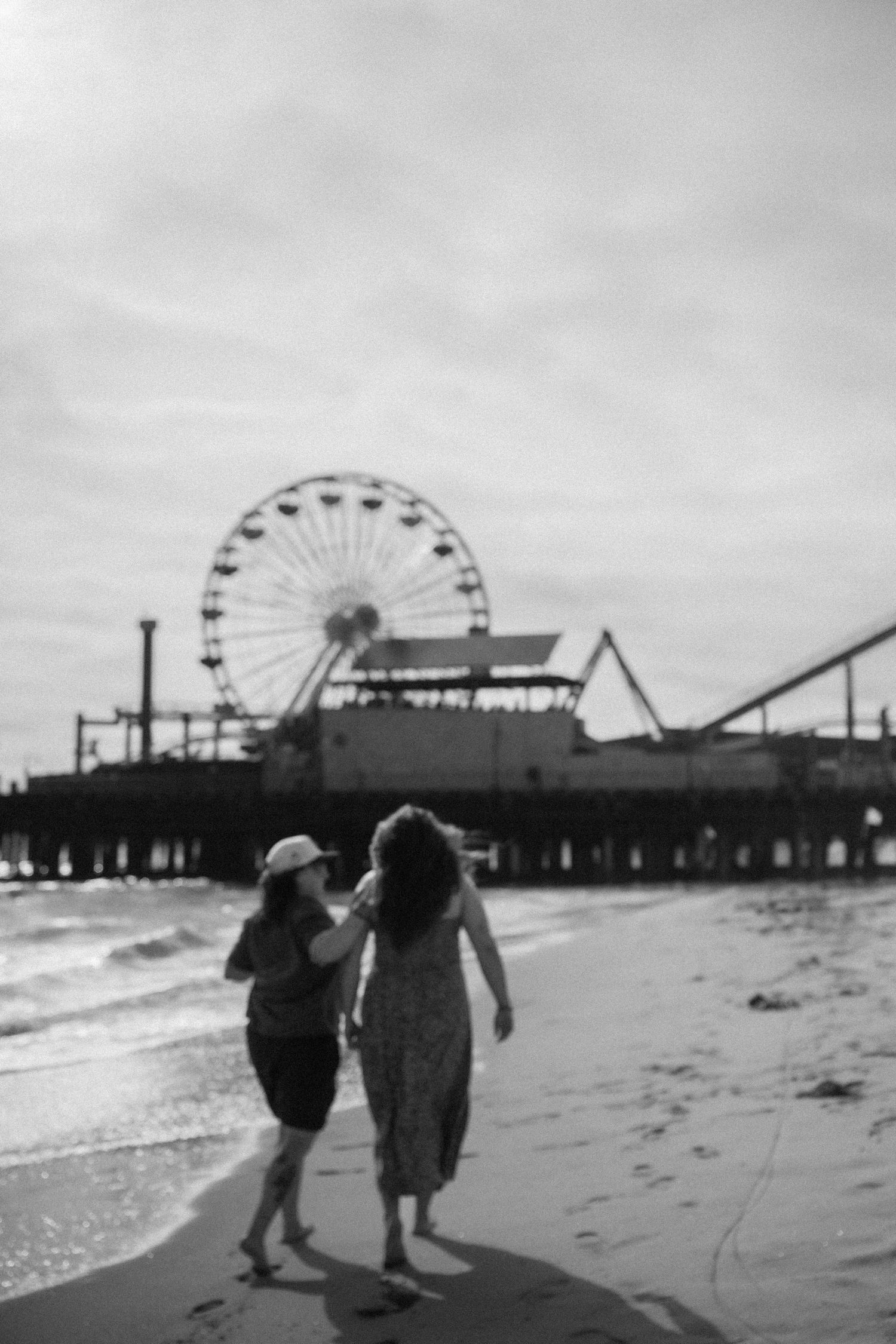 A black and white photo of two people walking near a pier with a Ferris wheel silhouette.