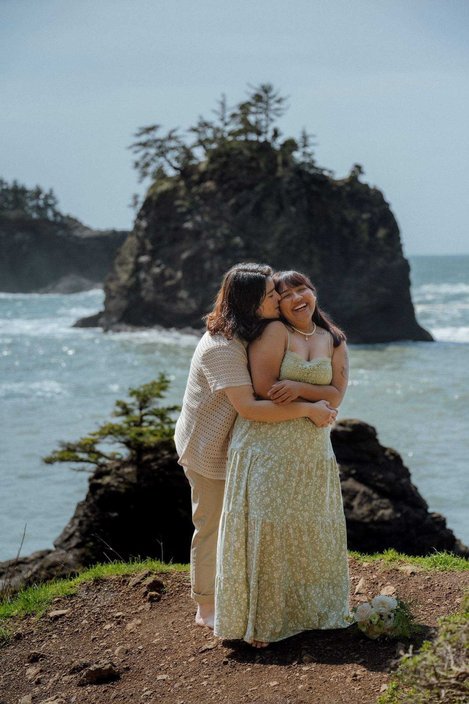 A couple shares a tender moment on a cliff overlooking dramatic coastal rock formations.