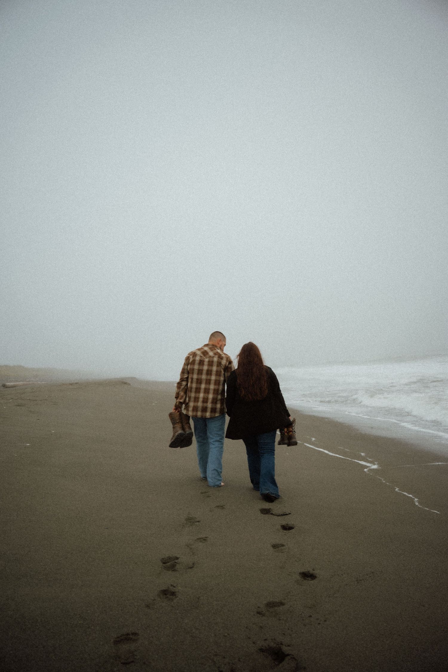 Two people walk together on a foggy beach leaving footprints in the wet sand.
