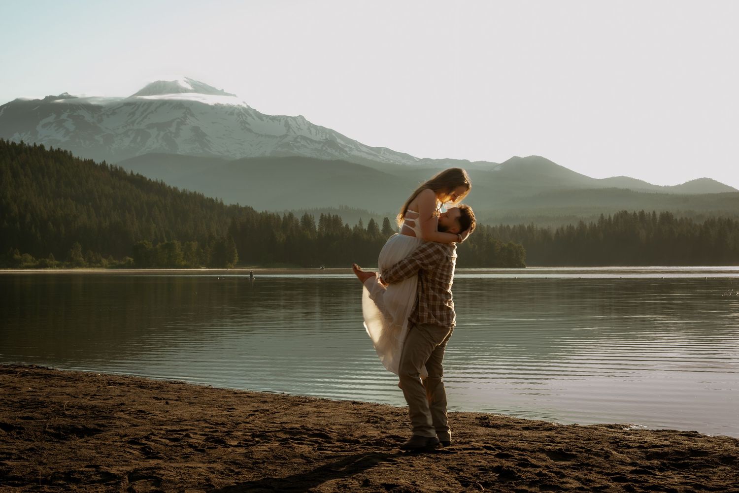 Couple embracing on a beach with mountains and lake in the misty background.
