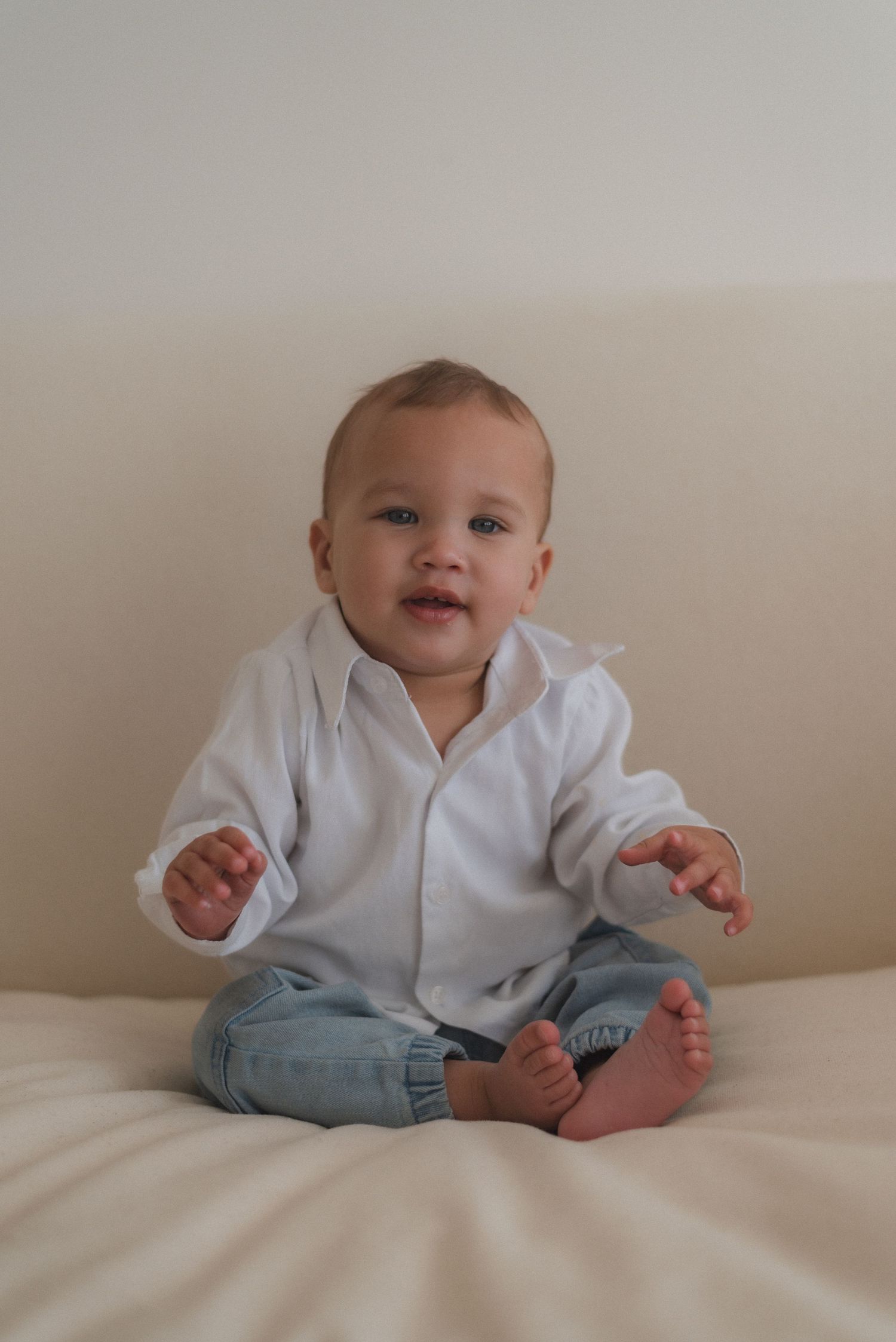 Baby in white button-up shirt and jeans sits on white bedding while playing.