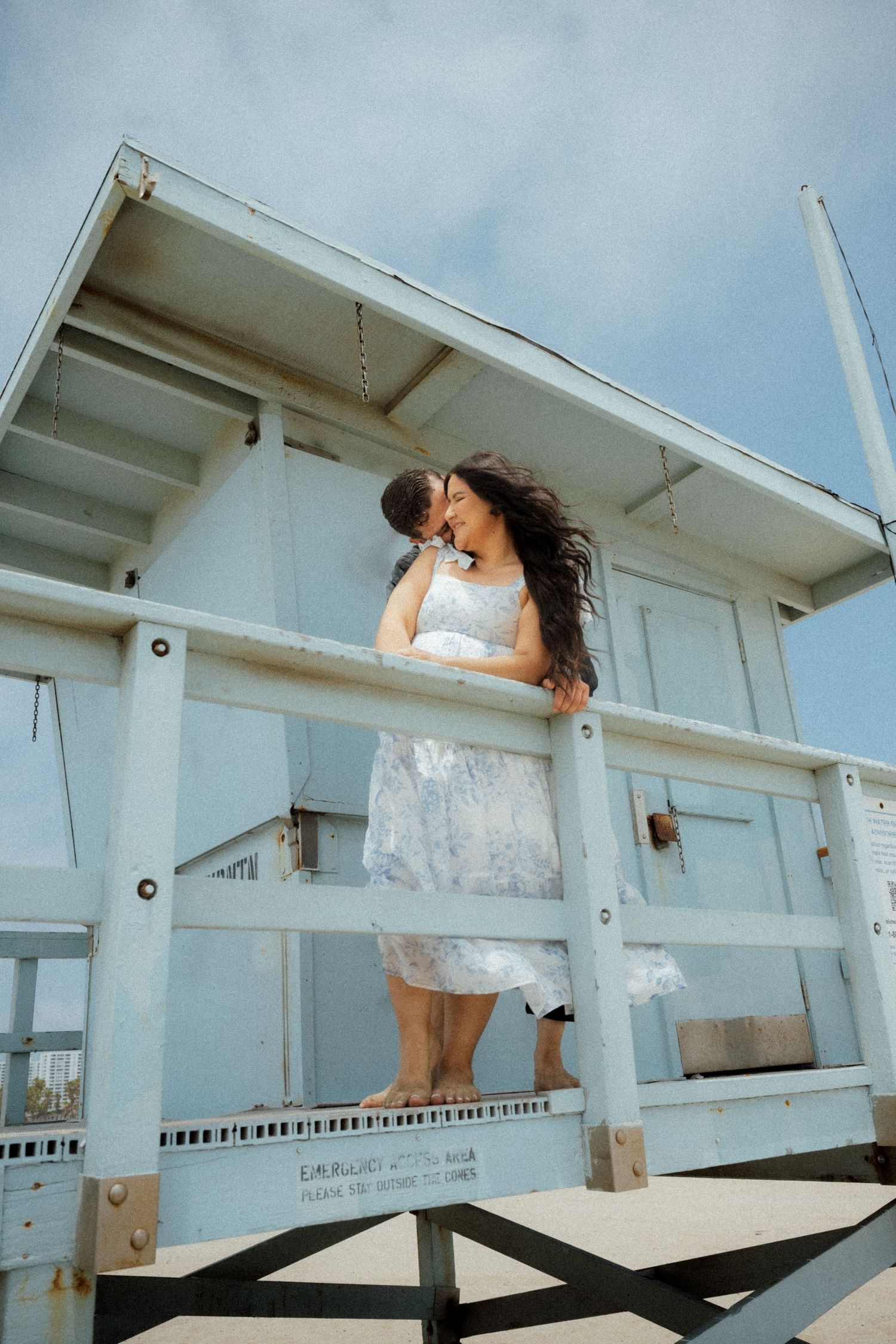A person in a flowy dress stands on a white lifeguard tower overlooking the beach.