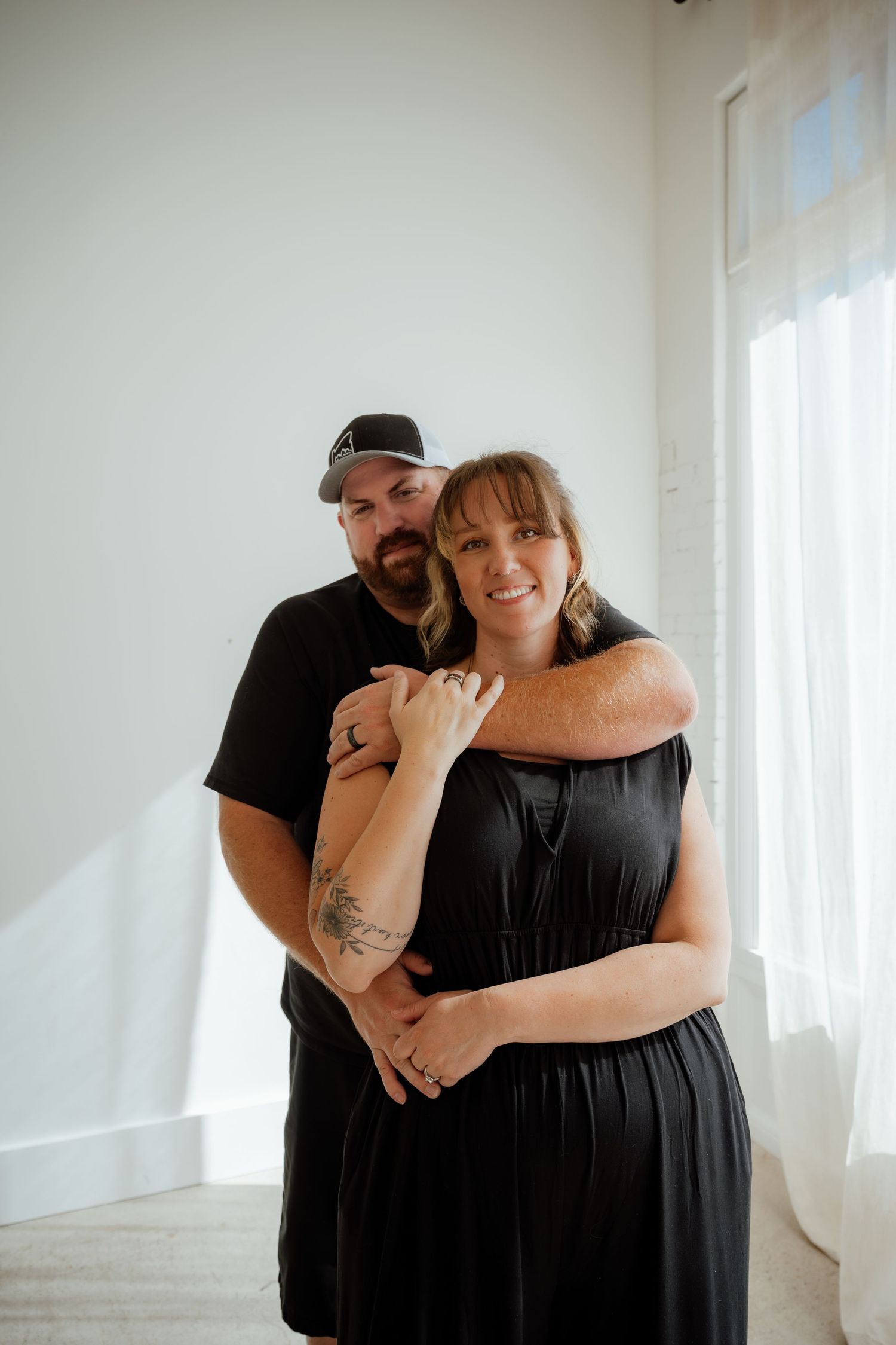 A couple poses together against a white wall in casual black outfits.