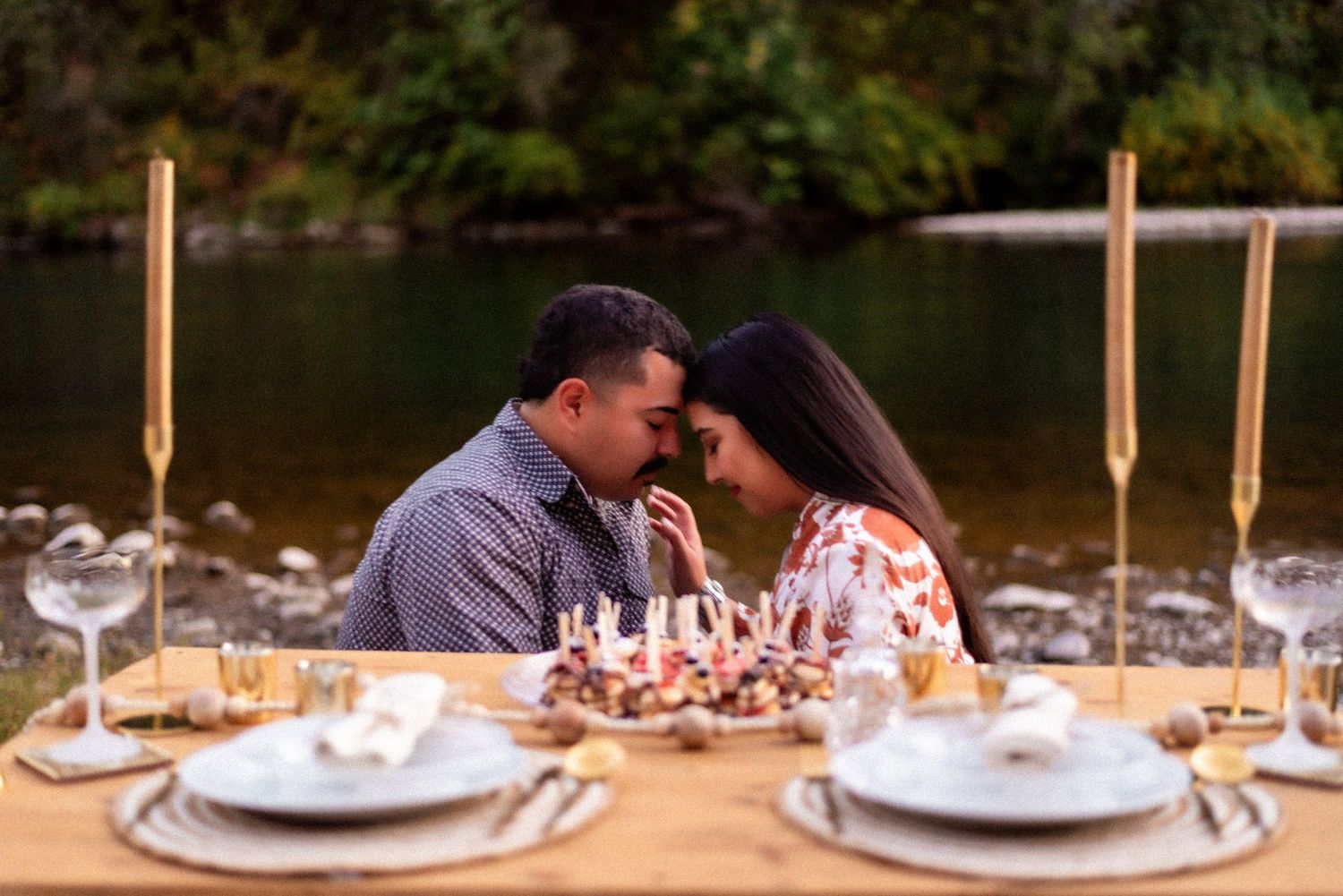 A romantic dinner setup by a lake with place settings and glowing candles at sunset.