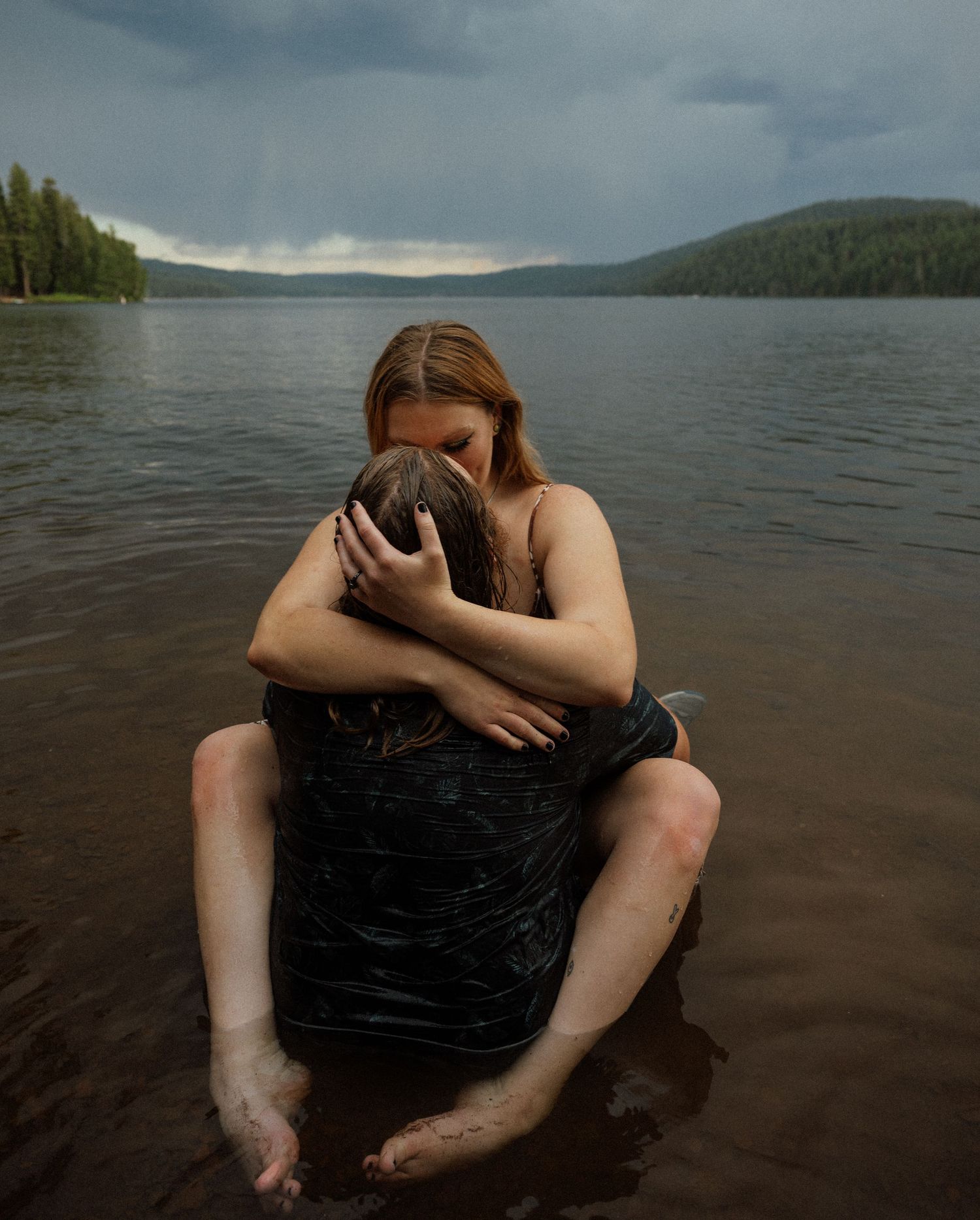 Couple holding each other while sitting in shallow lake water during an overcast day.