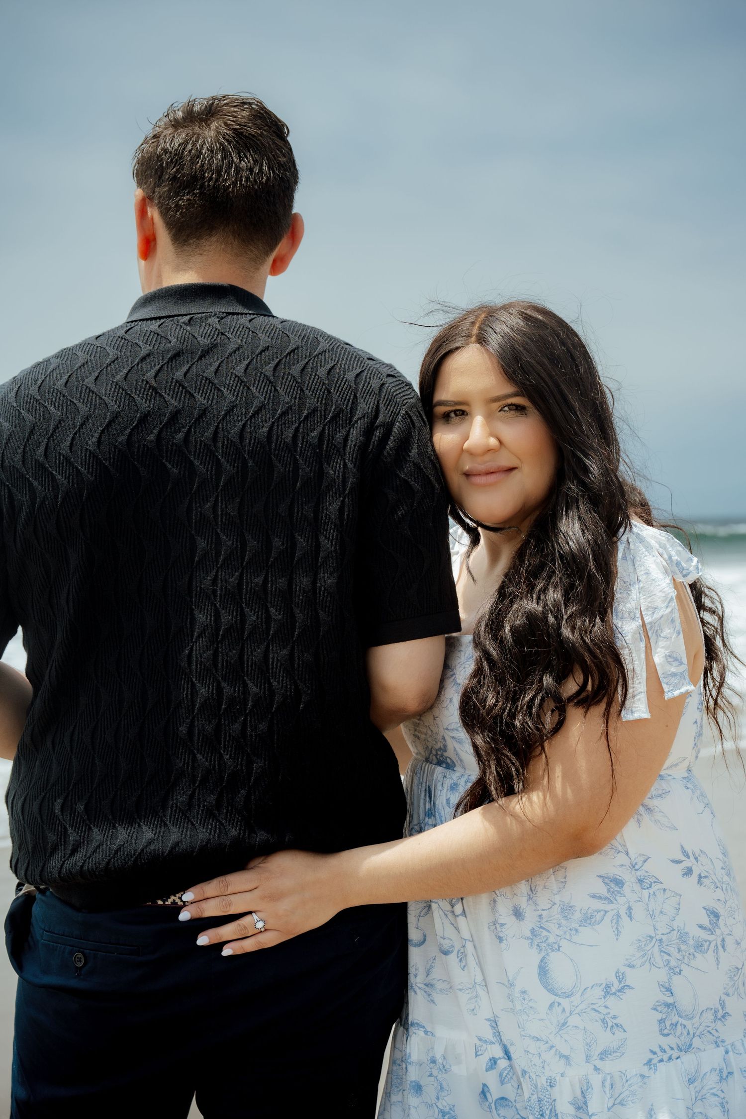 A couple poses together on a windy beach with ocean waves behind them.