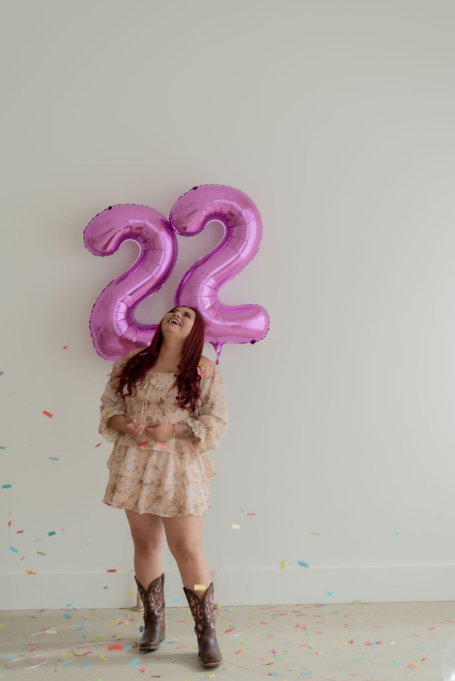 Person in a peach dress poses playfully with large pink number balloons and scattered confetti.