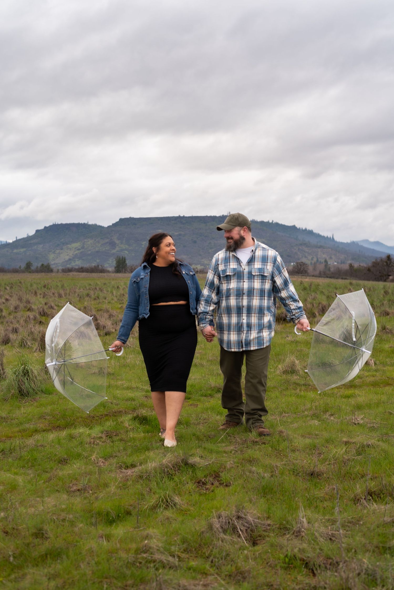 A couple shares a playful moment with umbrellas in a grassy field with mountains in the background.