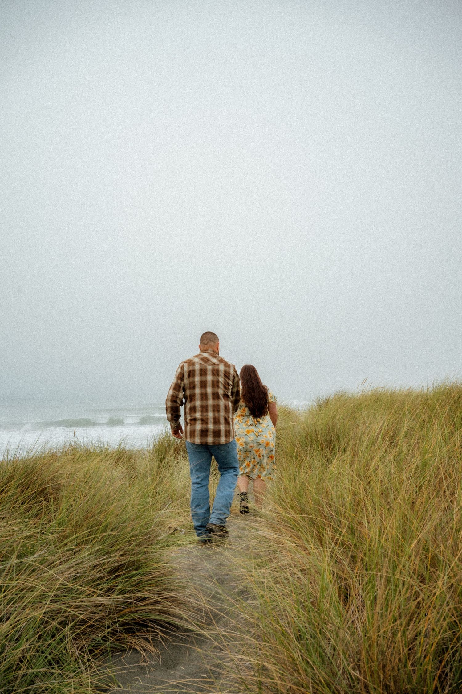 Two people walk along a coastal path through tall beach grass on an overcast day.
