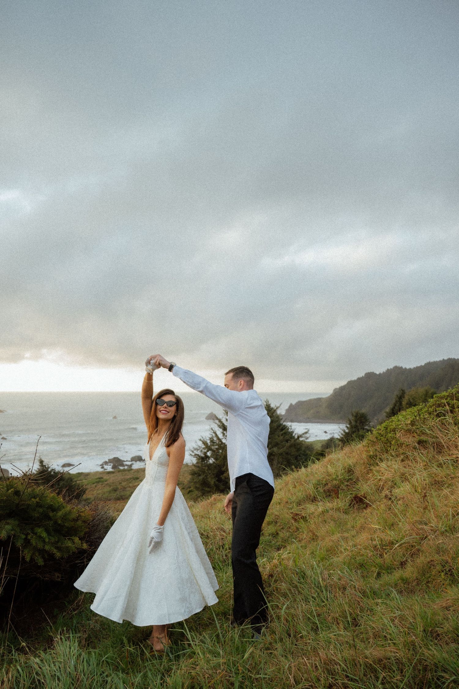 A couple dances together on a grassy coastal cliff with dramatic cloudy skies.