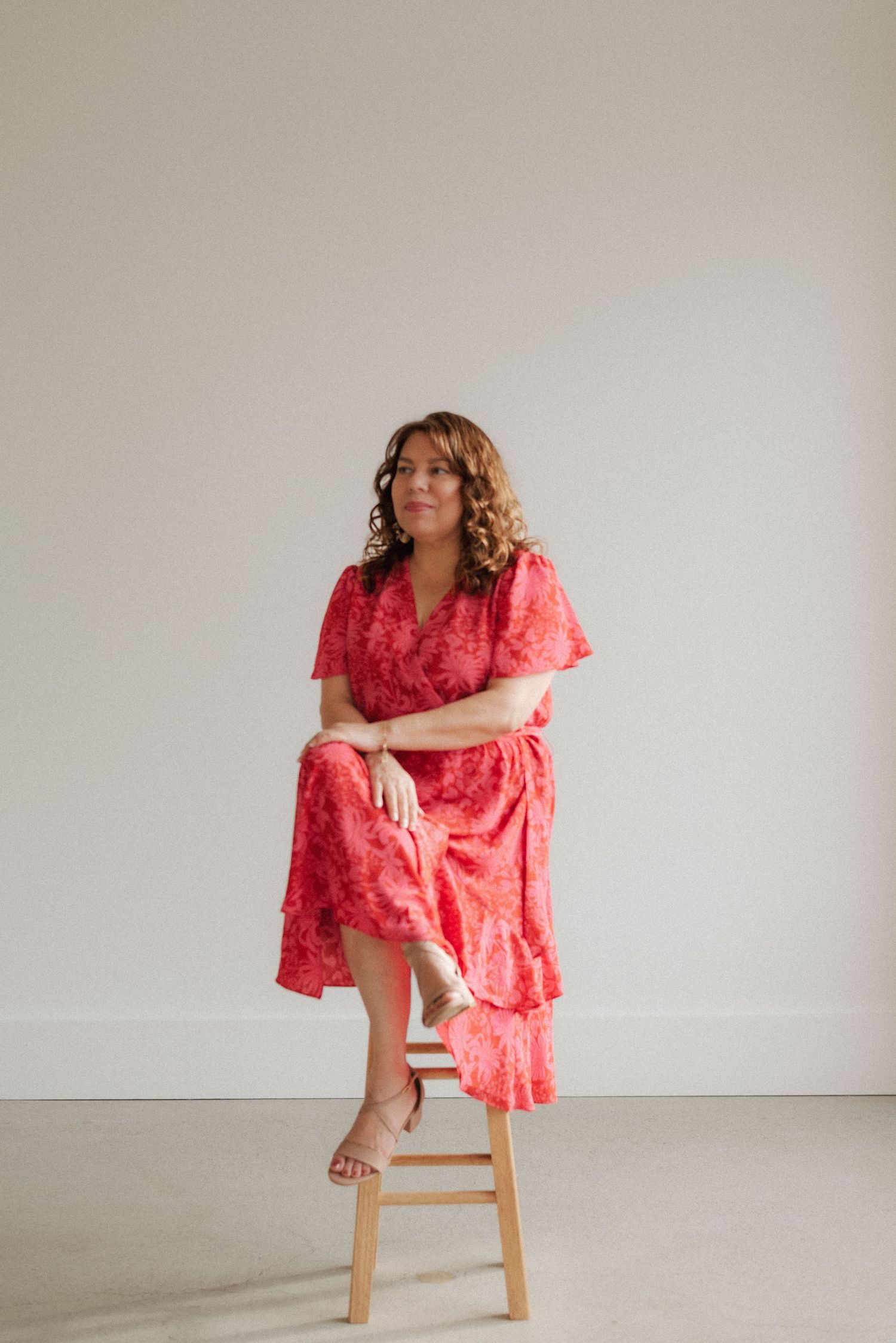 Fashion portrait of woman in red floral dress seated on wooden stool.
