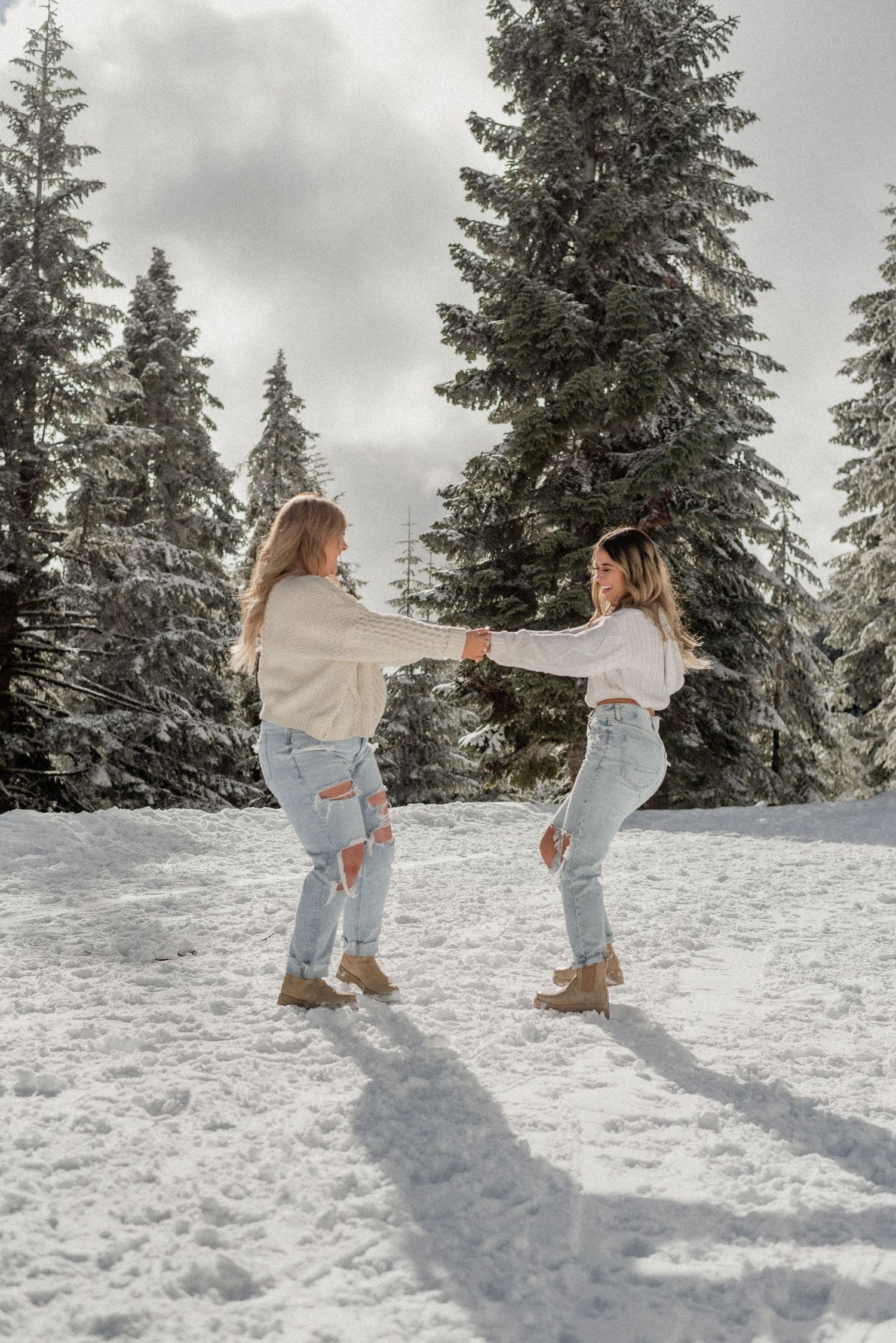 Two people playing in snow wearing matching cream sweaters and jeans against snowy pine trees.