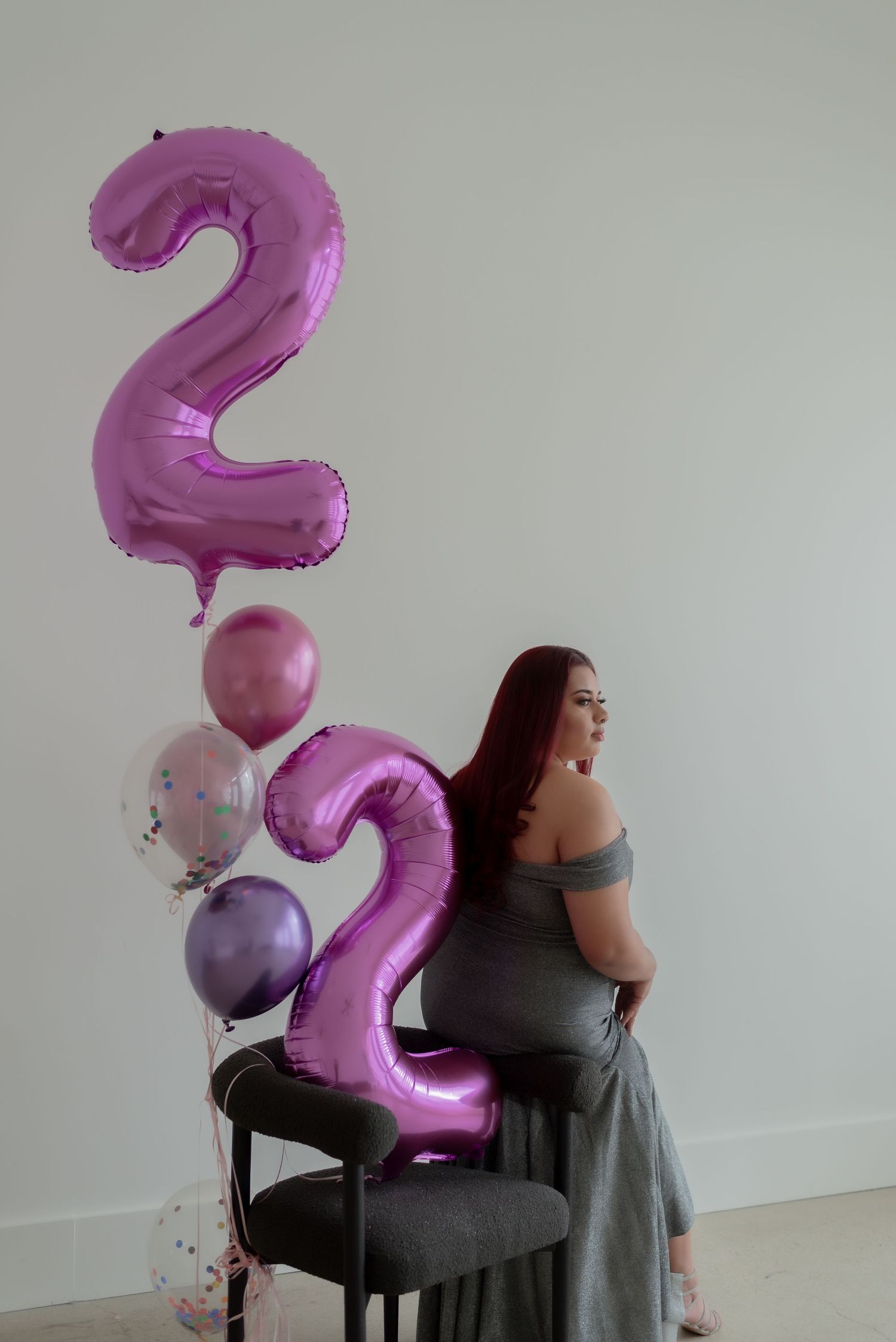 Woman seated with large pink number 2 balloon in minimalist studio setting.
