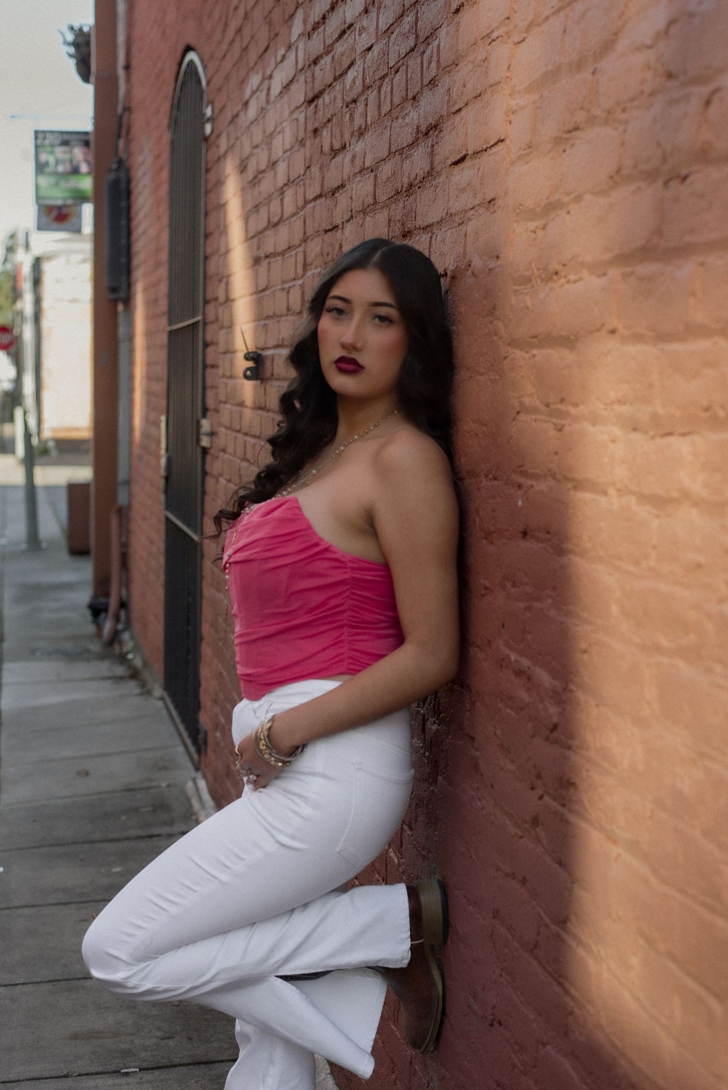 Fashion portrait with pink top and white pants against textured brick wall.