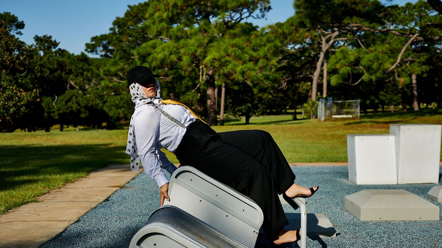 Woman in a white hijab and black pants leans back on an outdoor fitness machine in a sunny park surrounded by tall pine trees.