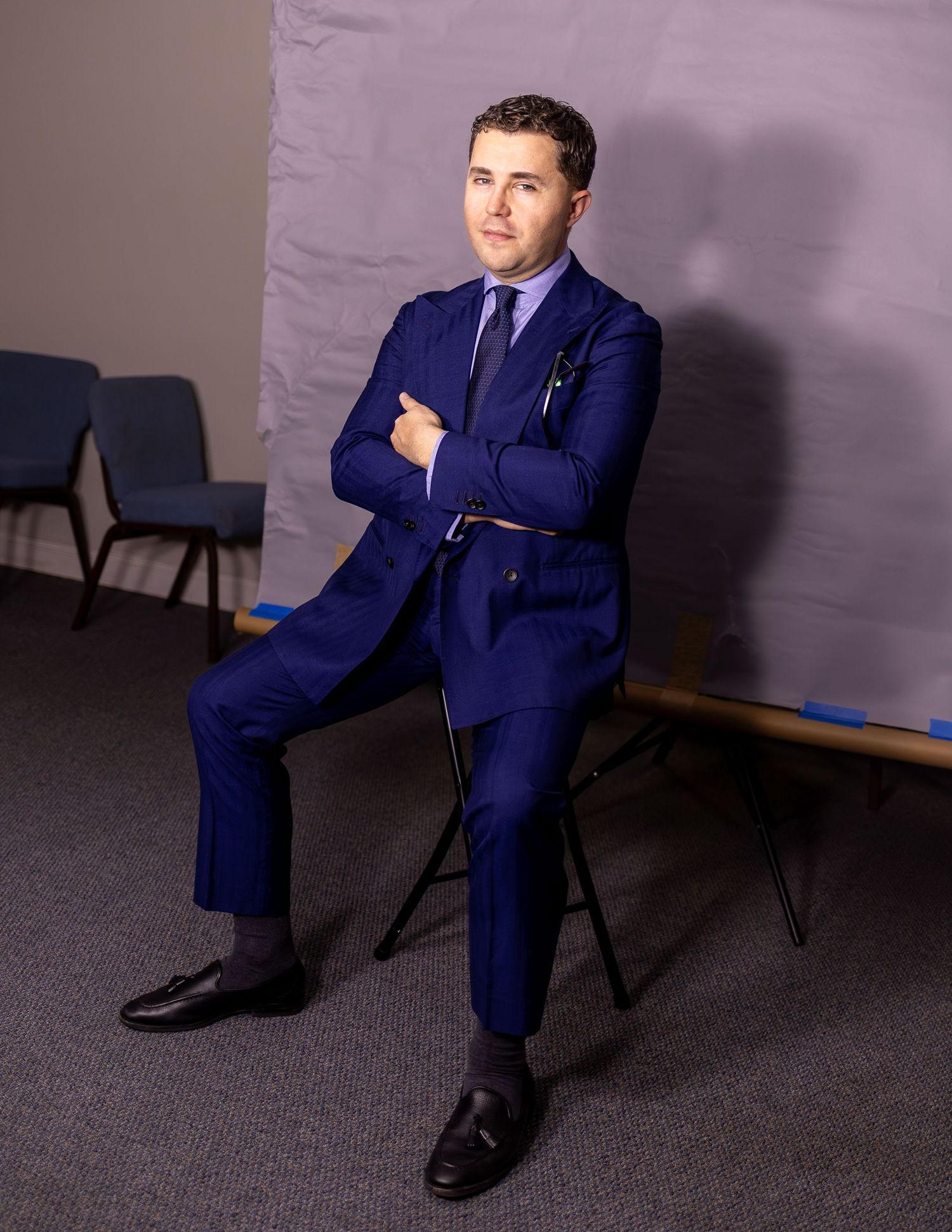 Man in navy blue double-breasted suit sitting cross-armed on stool in studio.