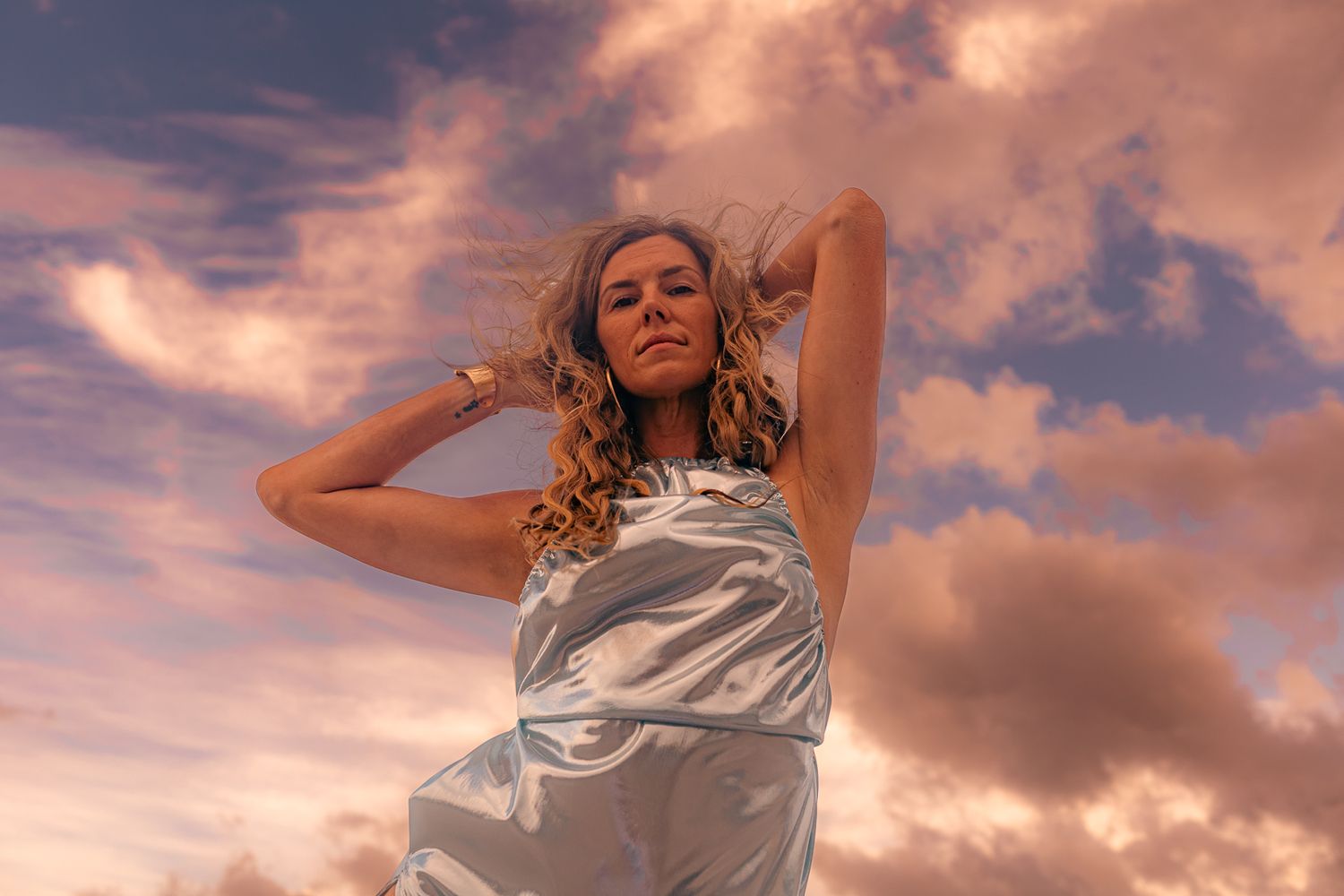 Woman in silver dress posing against dramatic cloudy sunset sky, hair blowing.