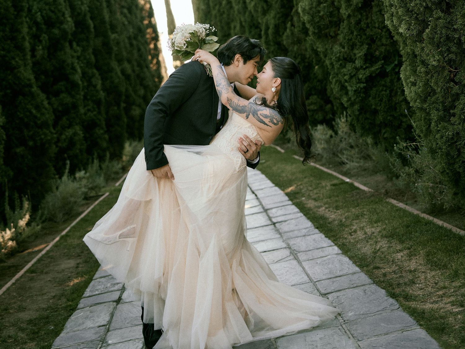 Groom dipping bride on a stone path surrounded by tall green cypress trees.